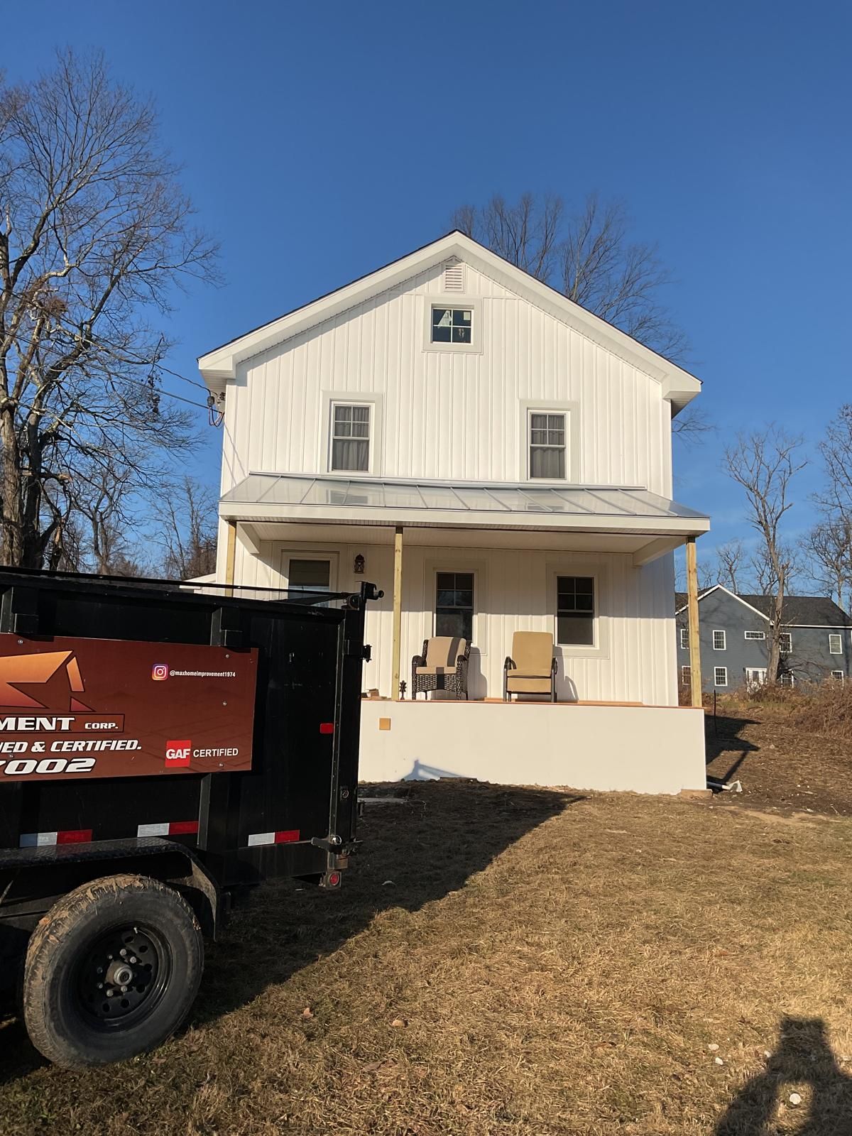 A two-story white farmhouse with a front porch and metal roof under a blue sky, partially obscured by a dump trailer.