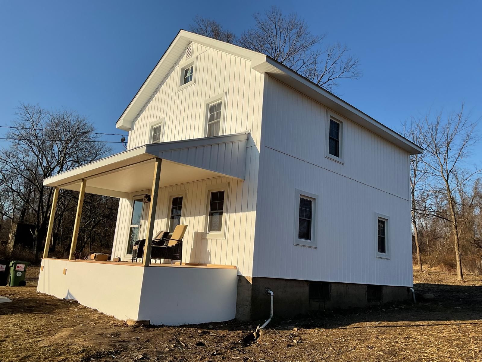 A white, two-story house with a front porch, located in a rural, sunlit setting under a clear blue sky.
