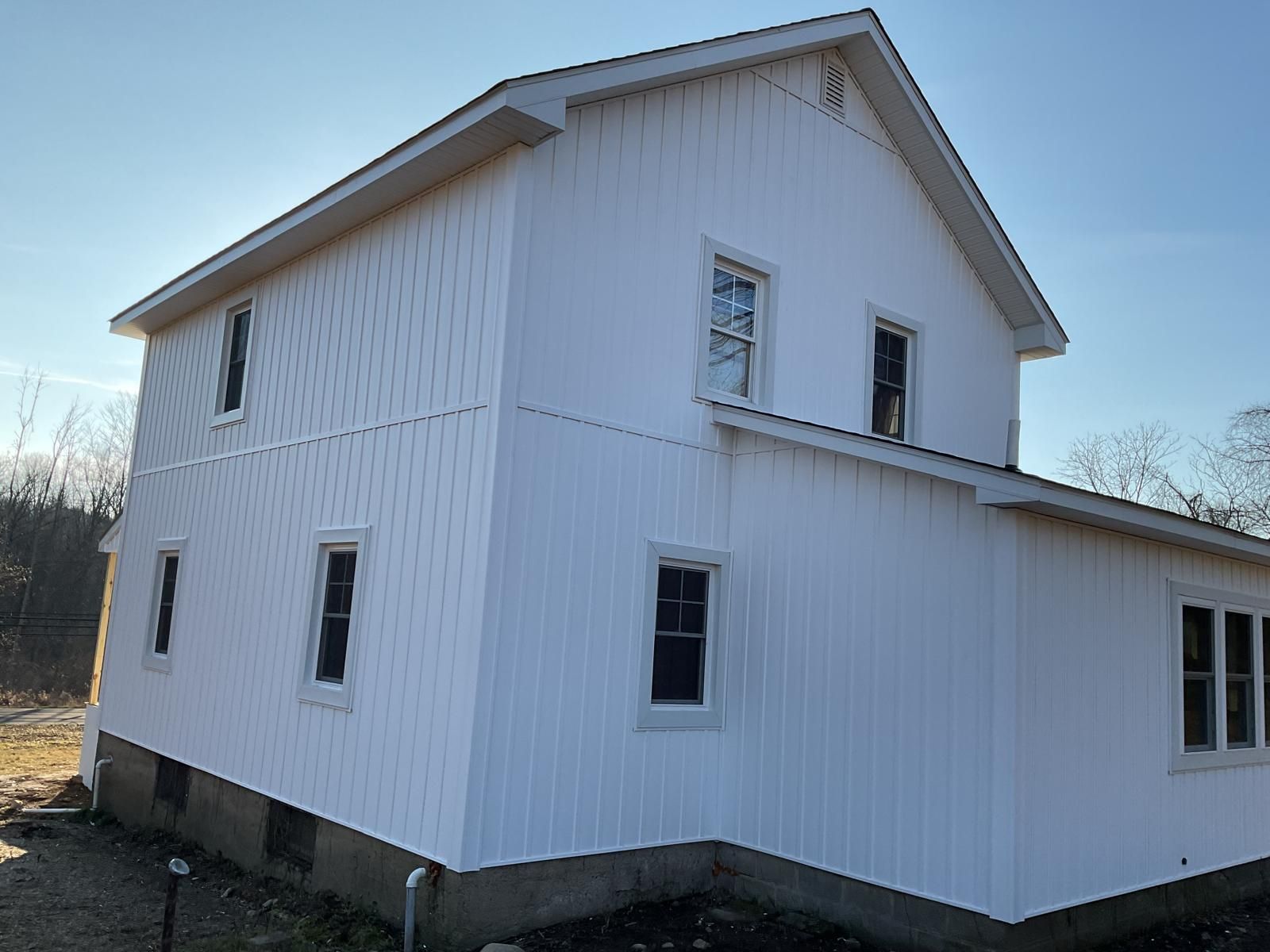 A two-story house with vertical white wood siding and multiple windows, viewed from an angle against a clear blue sky.