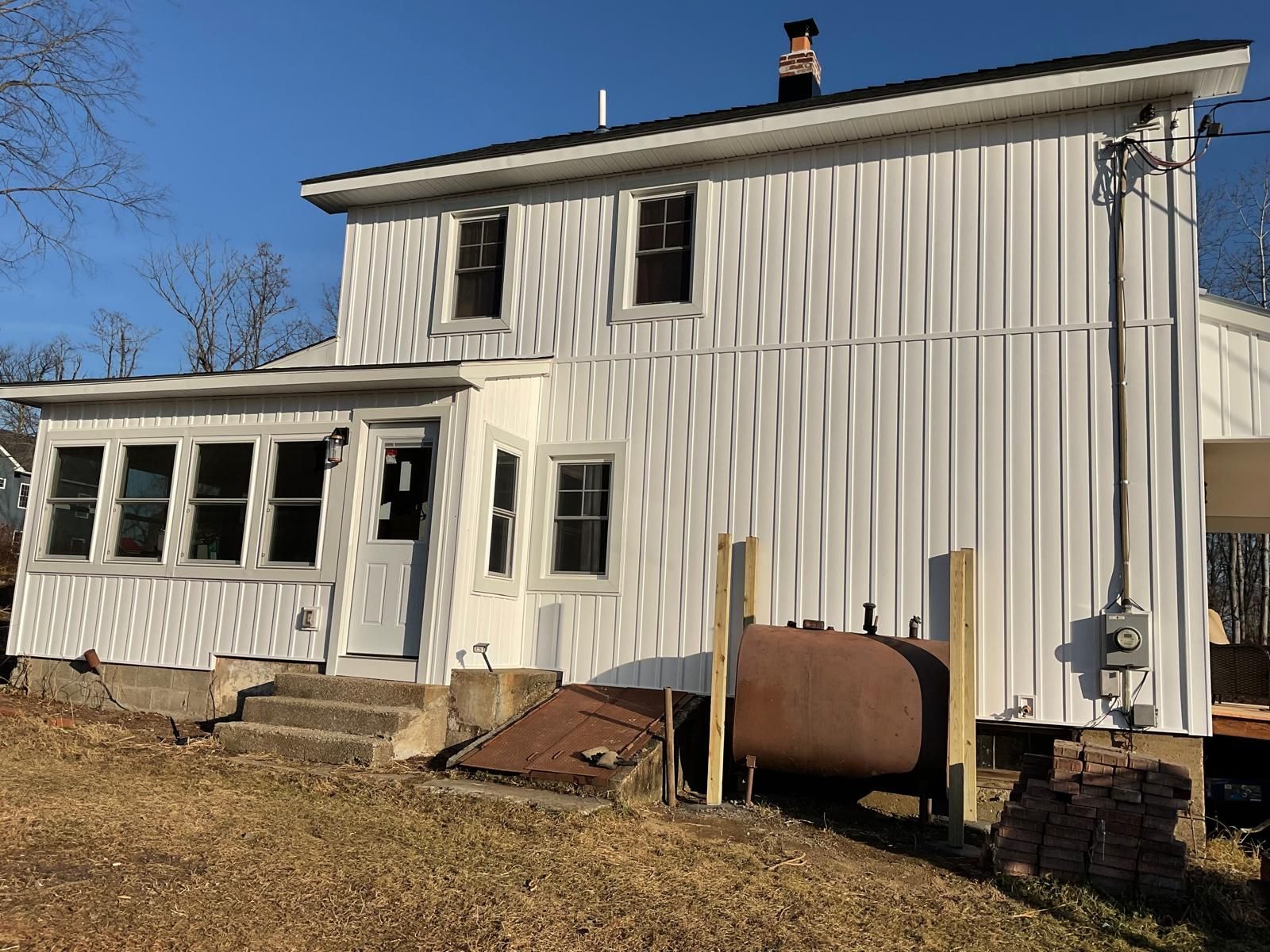 A two-story white house with vertical siding, a porch, and a brown fuel tank in the yard under a clear blue sky.