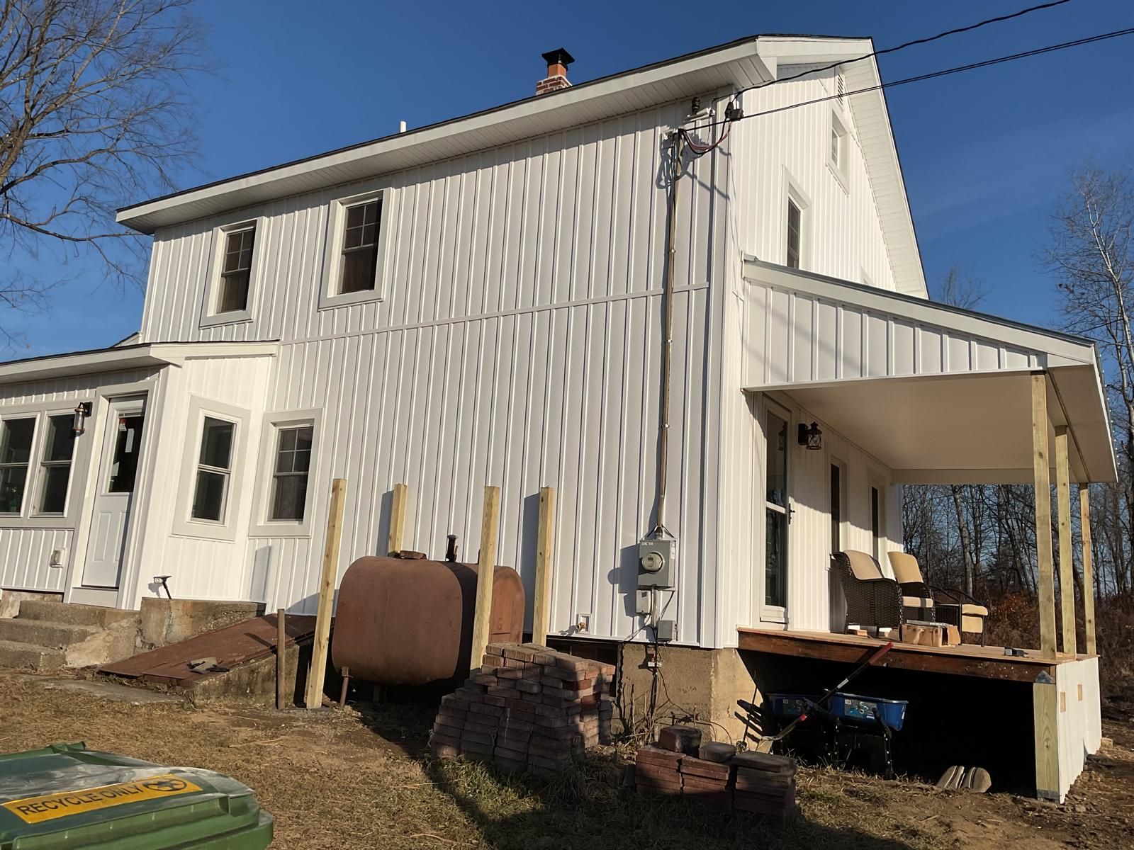 A white two-story house with board-and-batten siding, a rusty oil tank, and an unfinished porch on a sunny day.
