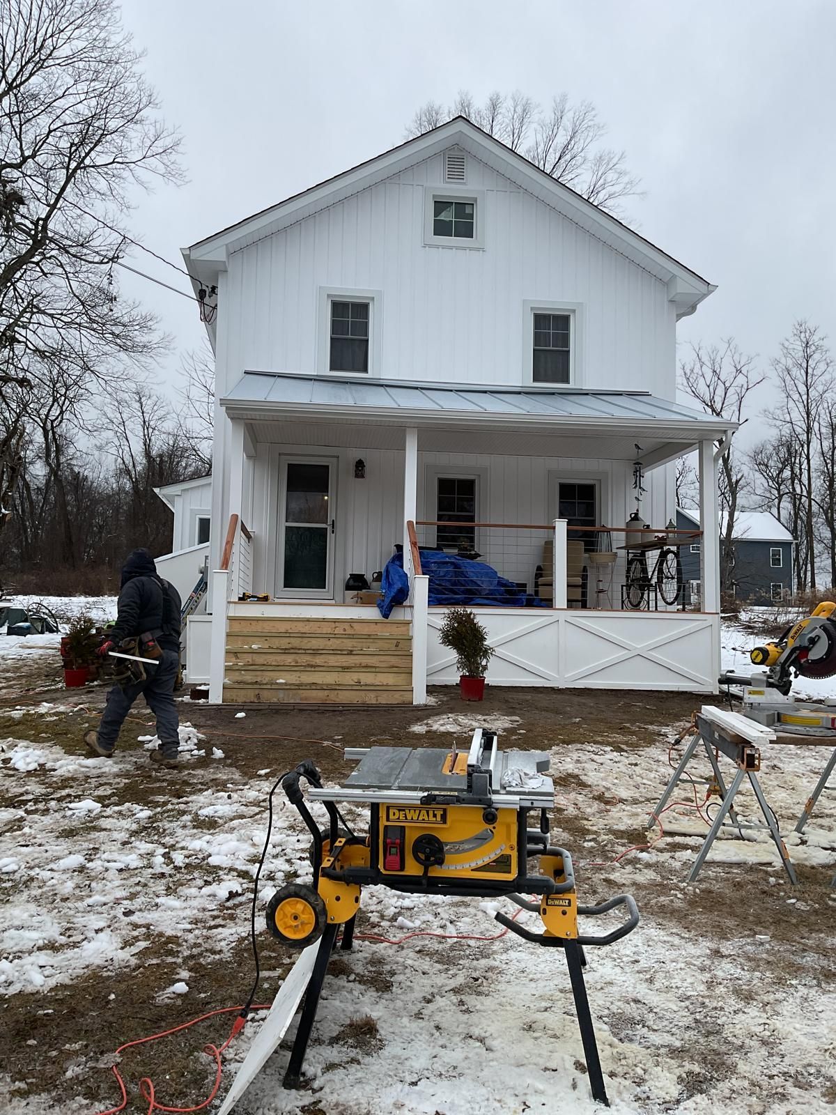 A person works on a snowy lawn in front of a white two-story house with a partially renovated wooden porch.