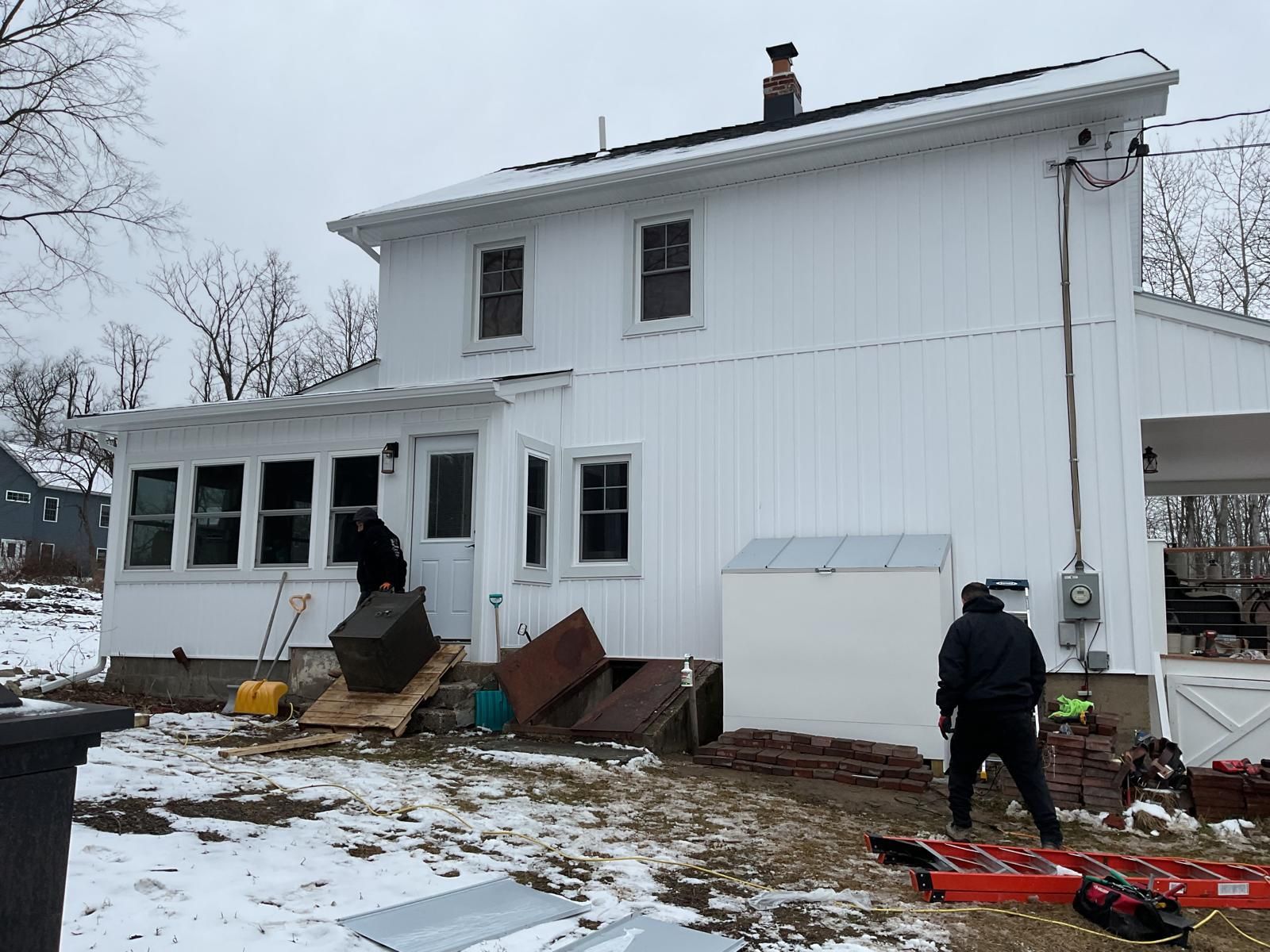 Two workers in dark clothing perform exterior maintenance on a white two-story house with snow on the ground.