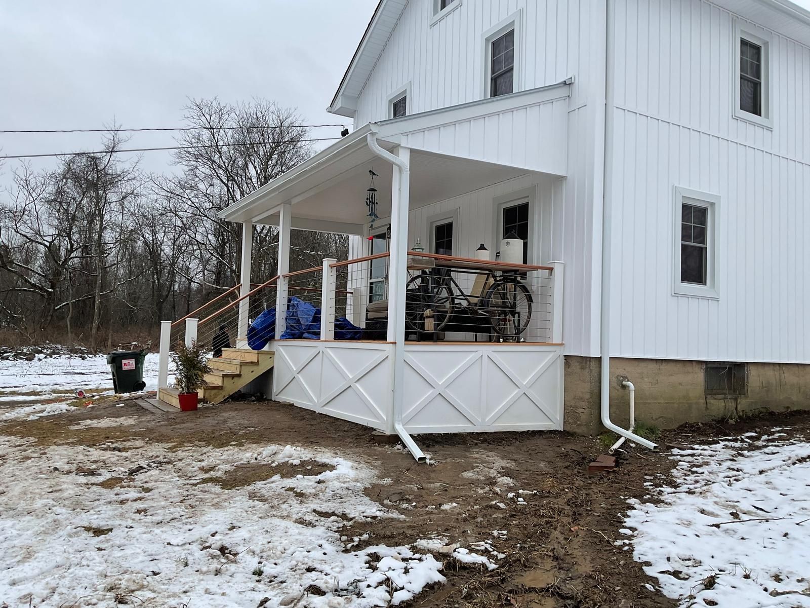 A white house with a covered porch featuring white lattice, stairs, and bicycles, set in a snowy landscape.