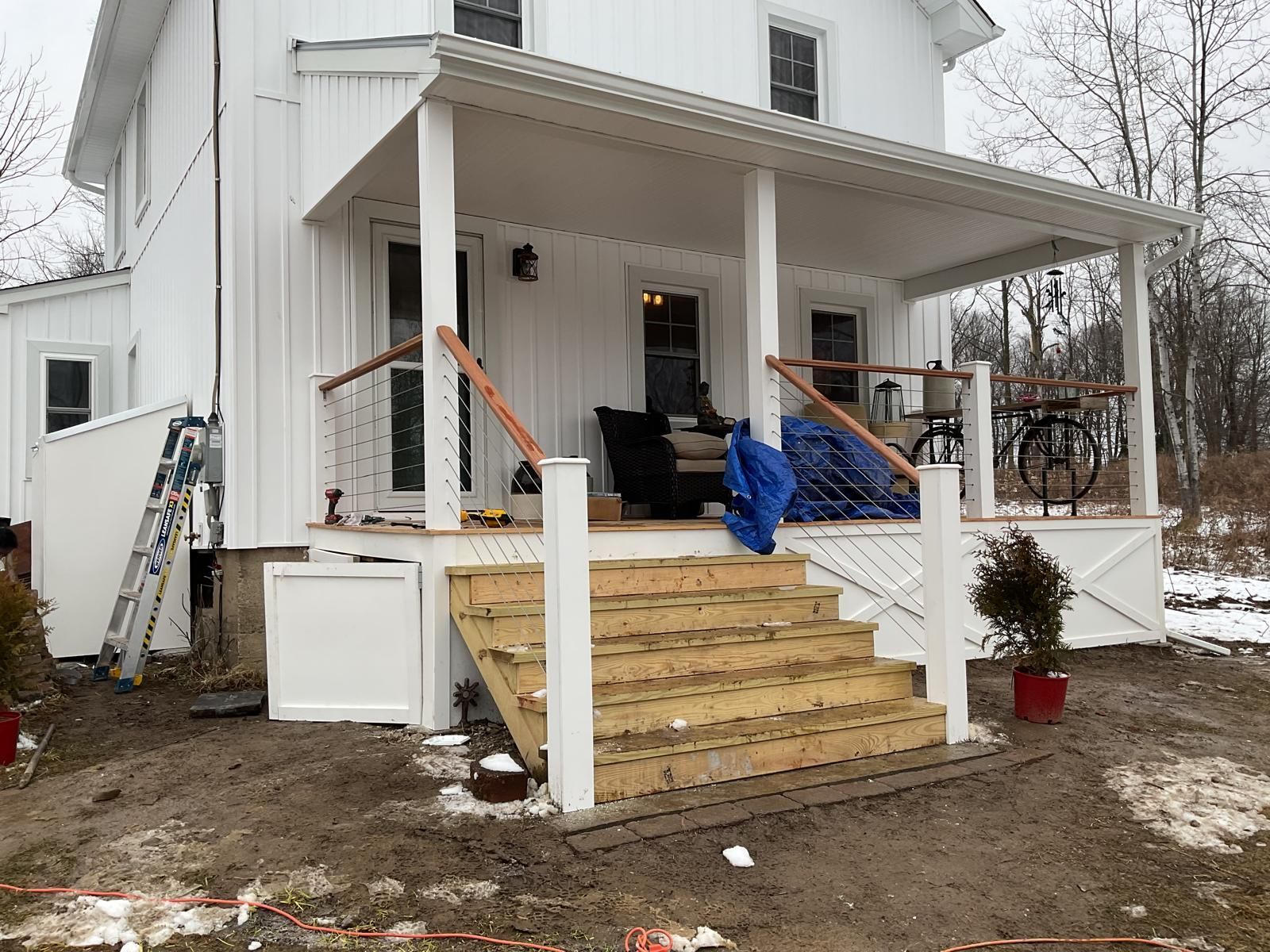 White farmhouse with a porch under construction, featuring new wooden stairs and cable railings.