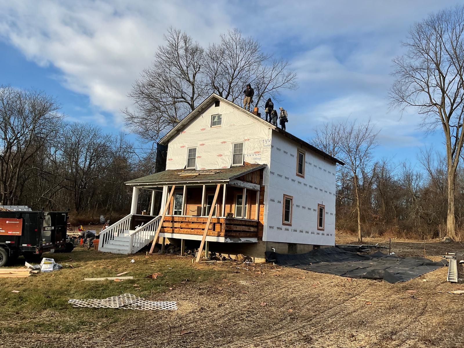 Workers stand on the roof of a two-story farmhouse undergoing exterior renovations in a rural setting.