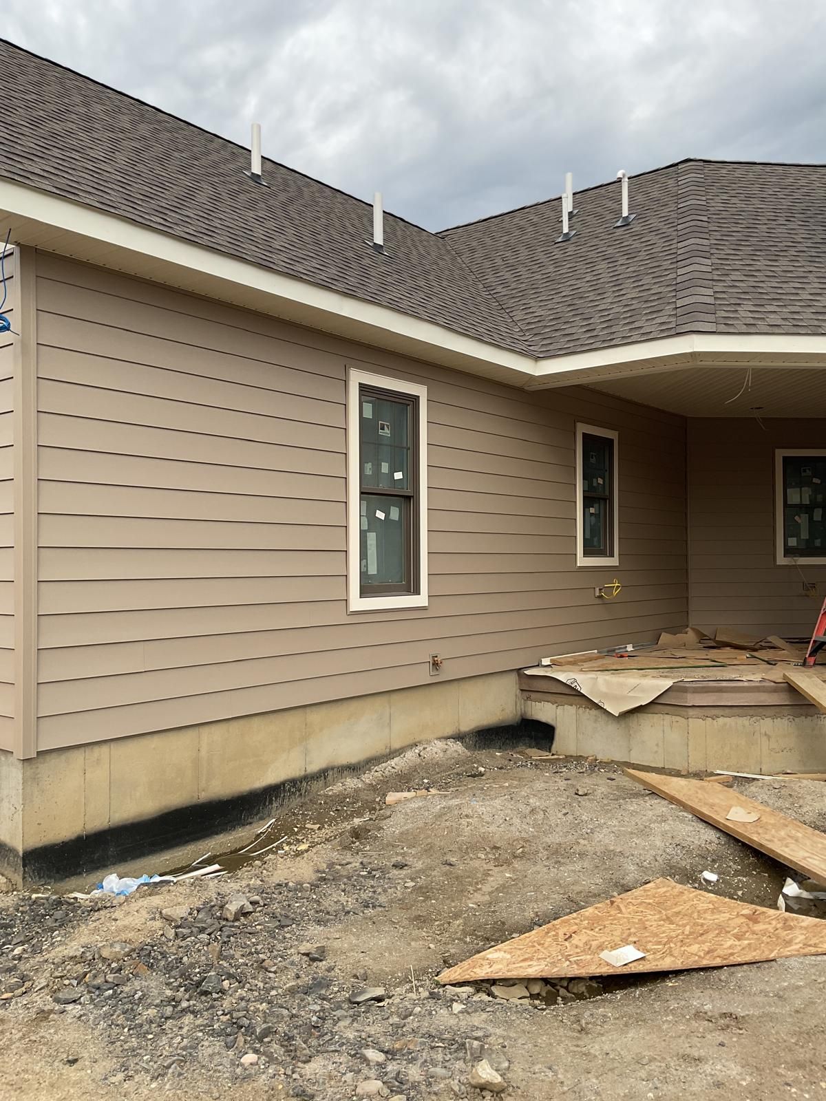Tan, wood-sided residential building under construction with a dark shingle roof and foundation exposed at ground level.