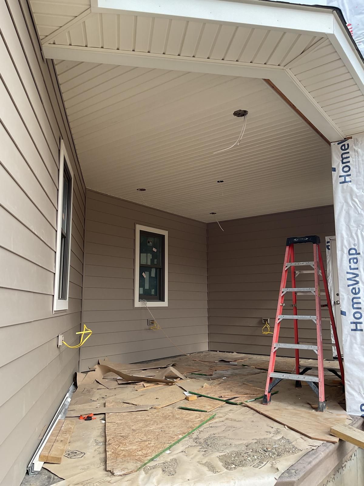 A partially finished covered porch with light brown siding, a red ladder, and an exposed ceiling on a construction site.