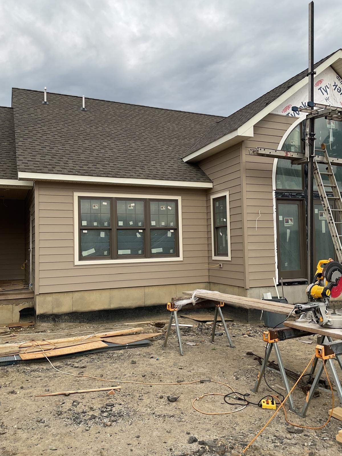 Exterior view of a house under construction with beige siding, a brown roof, large windows, and a workstation on site.