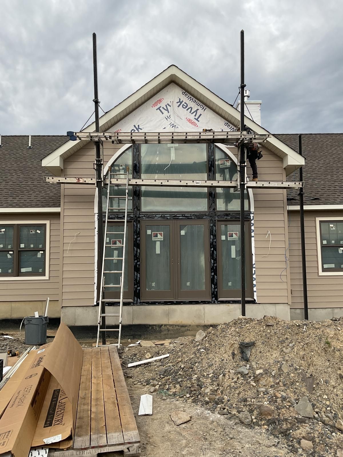 Construction workers on scaffolding installing a large window and door assembly on a tan house with a gable roof.