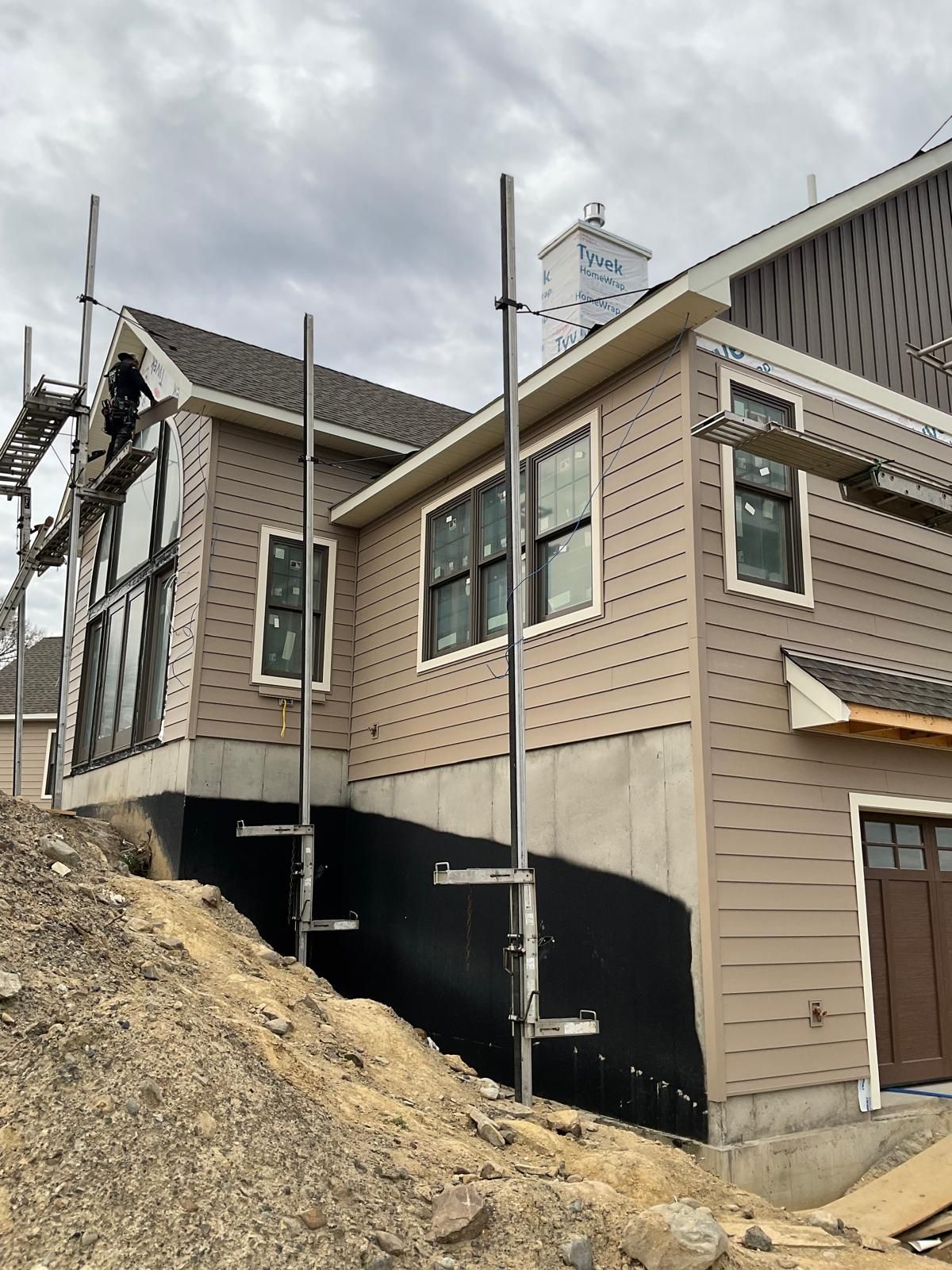 A house under construction with tan siding and a black waterproof foundation membrane, surrounded by scaffolding.