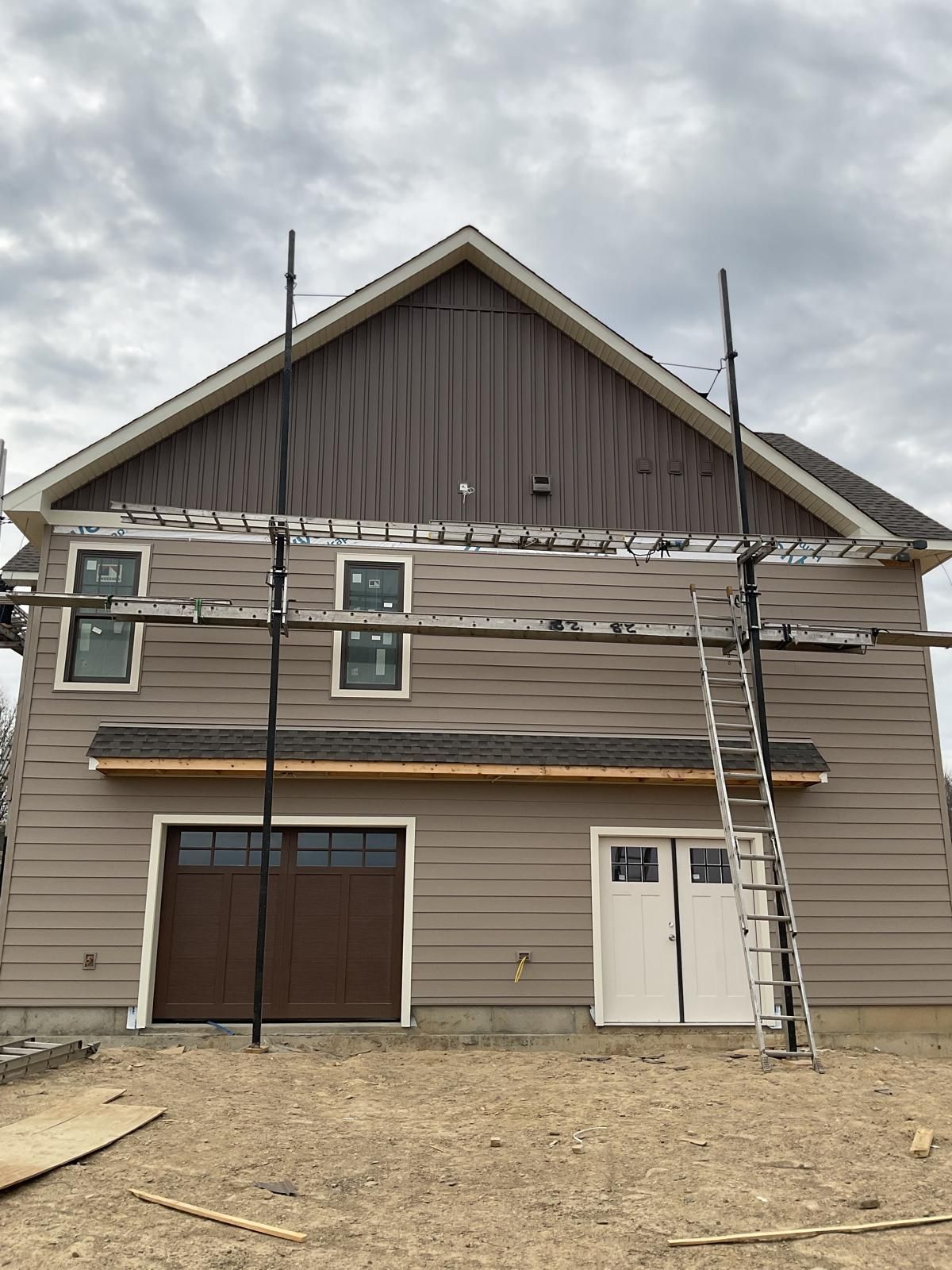 A two-story house under construction with tan horizontal siding, dark brown gable siding, and metal scaffolding.