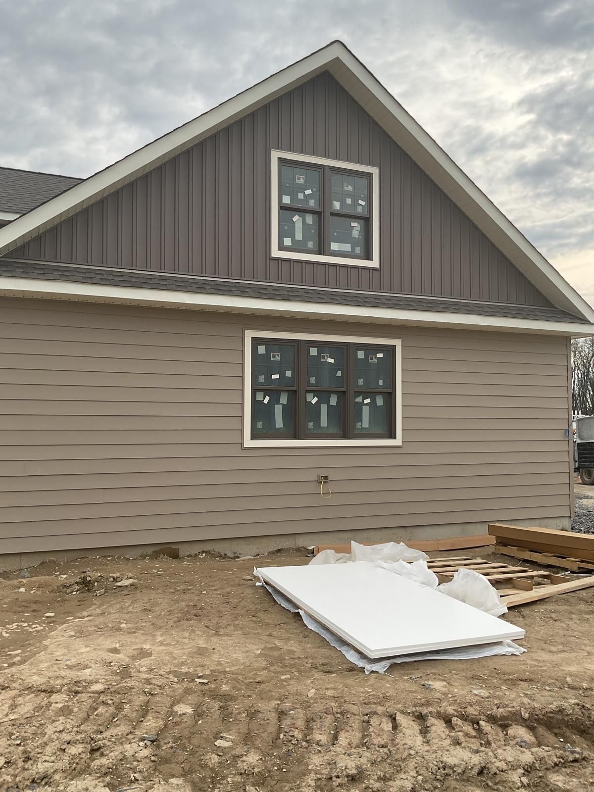 A newly constructed house exterior with light brown horizontal siding, dark vertical gable siding, and two black windows.