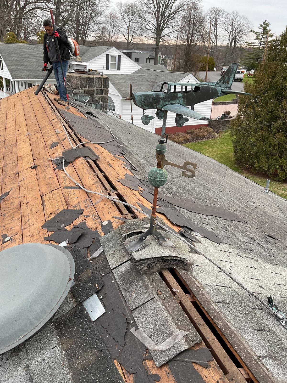 A person uses a leaf blower on a roof being renovated, featuring an airplane weather vane in the foreground.