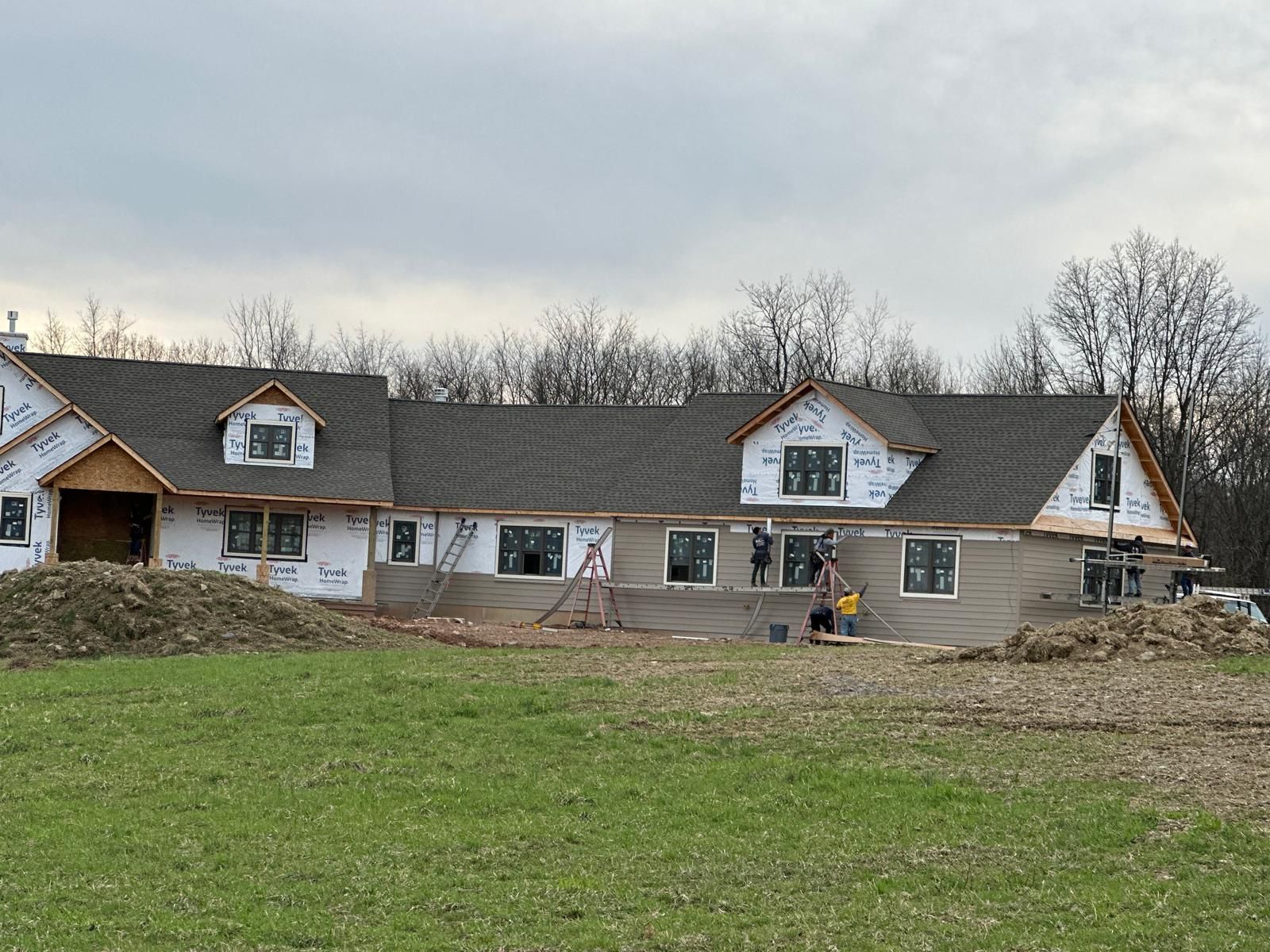 A house under construction with roof shingles, white house wrap, and workers standing in front of the exterior walls.