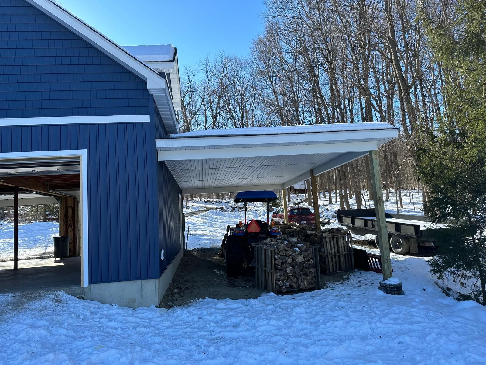 Blue-sided barn with an open lean-to shelter storing stacked firewood in a snowy, wooded outdoor setting.