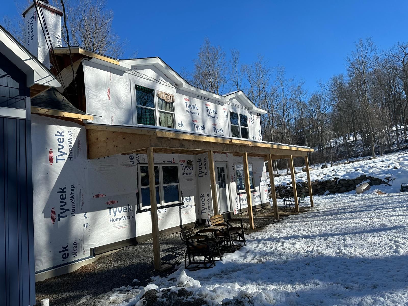 A house under renovation, covered in white Tyvek wrap, with a newly framed wooden porch in a snowy, wooded setting.