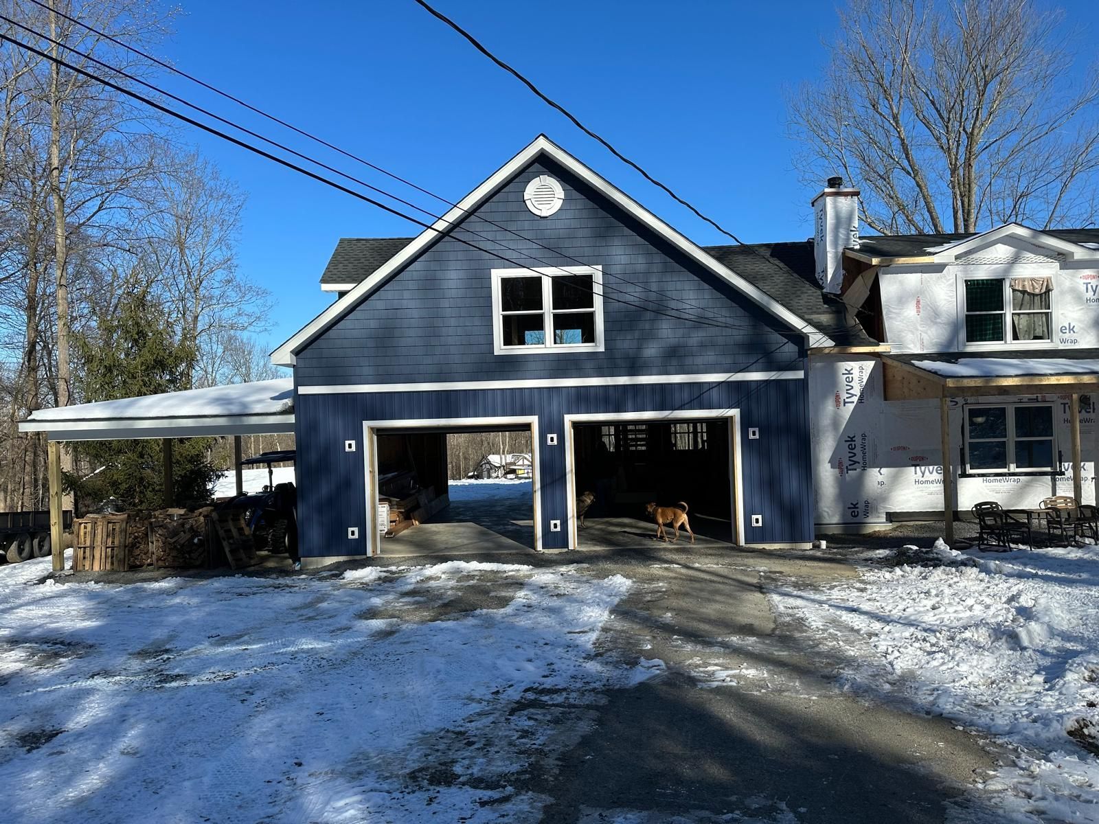A blue two-story detached garage with a covered side area and an attached white house, set in a snowy, wooded yard.