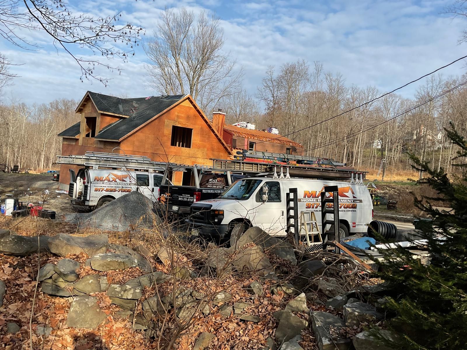 Two white utility vans parked in front of a partially constructed two-story house with a wooden frame on a wooded lot.