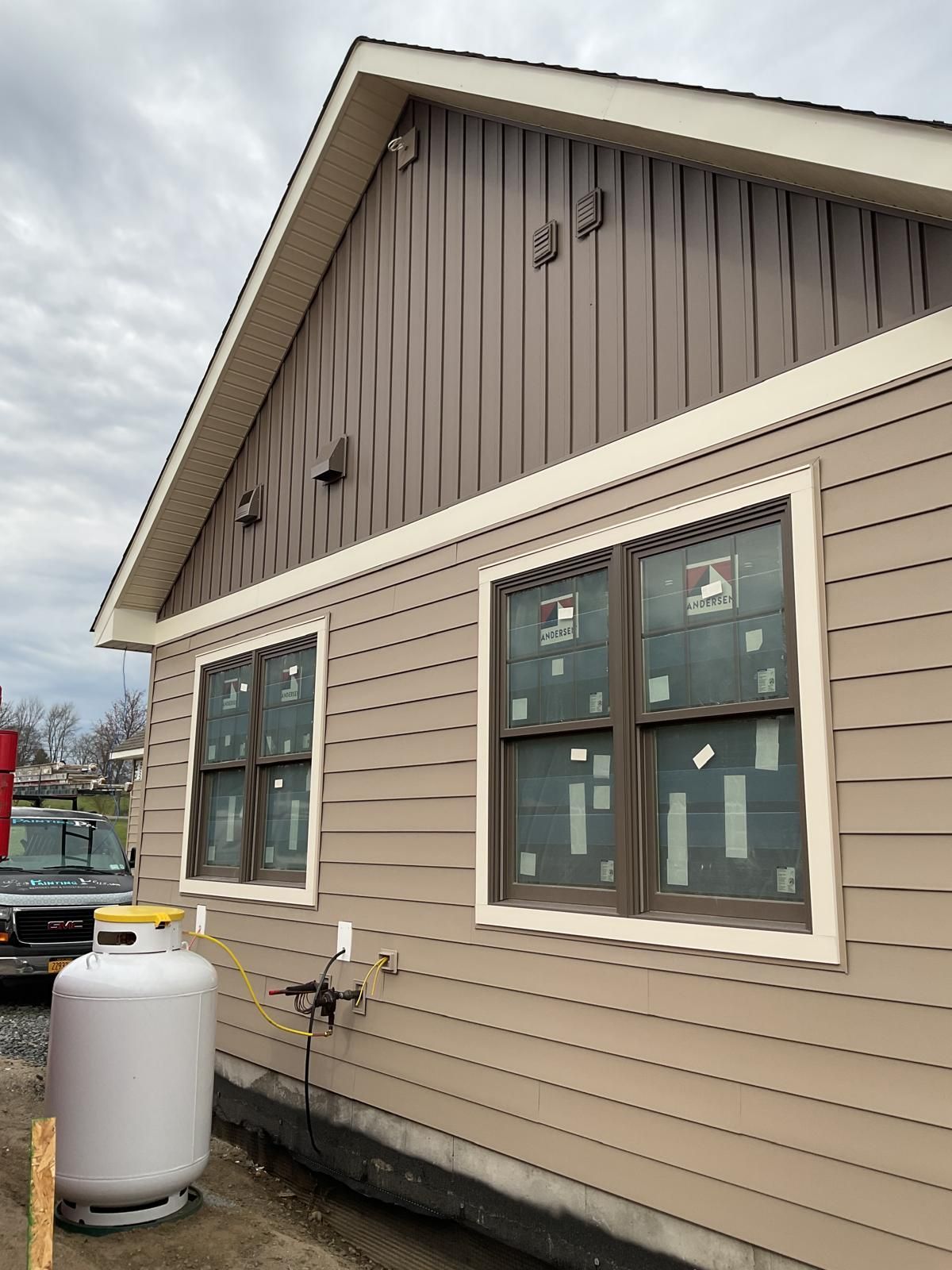 Exterior view of a tan-sided home under construction with a brown vertical-sided gable and a large propane tank.