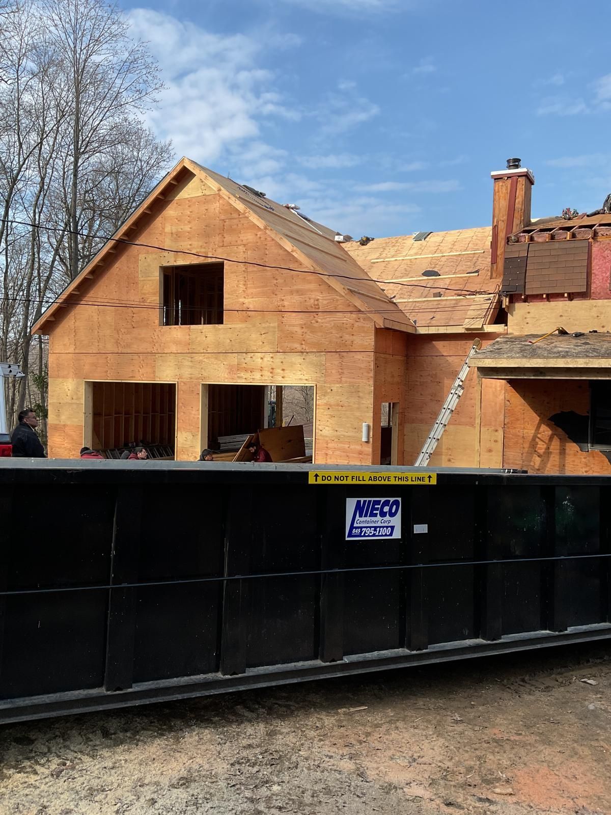 A partially framed house under construction with a large, black dumpster in the foreground under a blue sky.