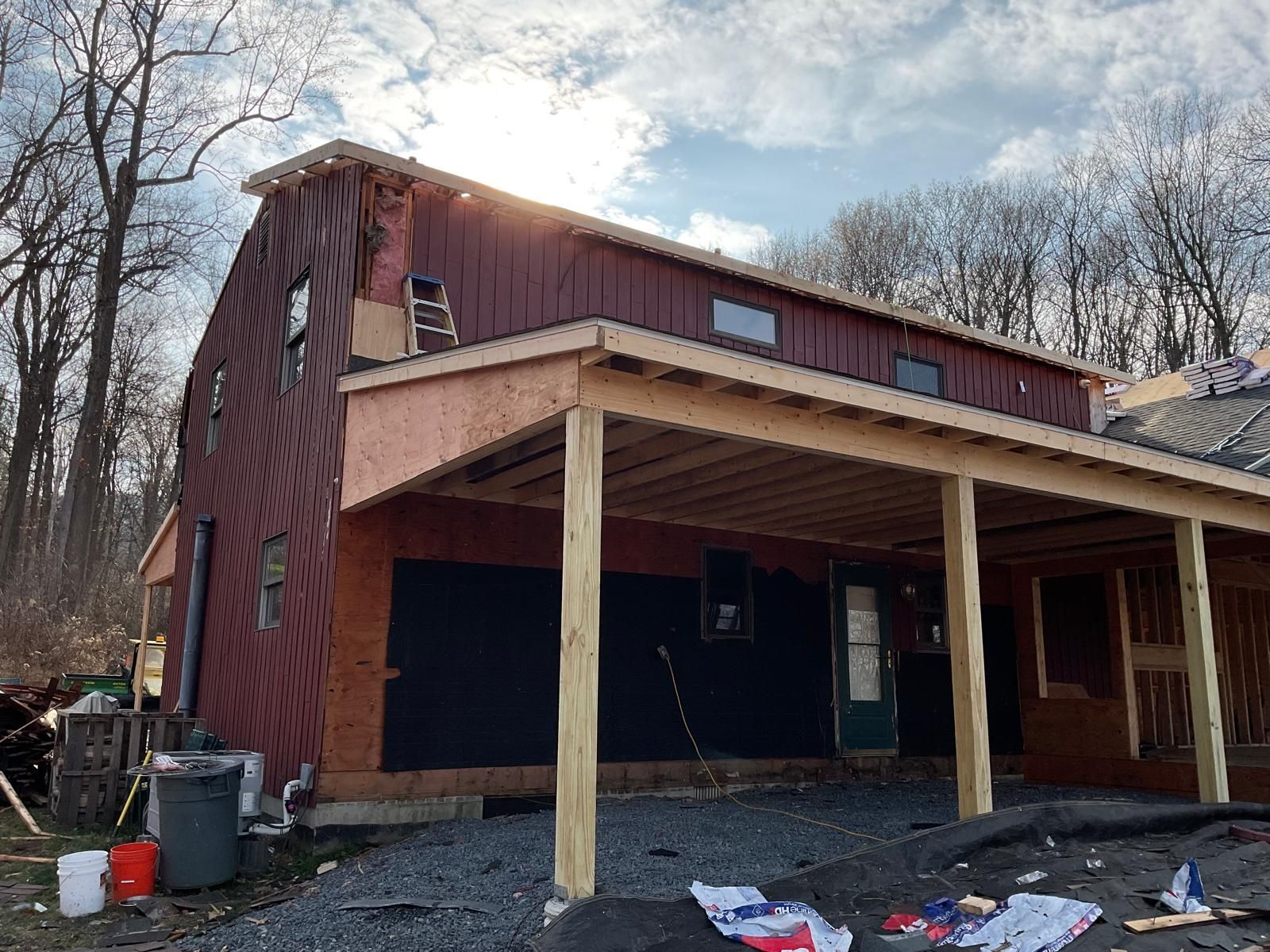 A partially constructed barn-style building with red siding and a wooden porch, set against a backdrop of bare trees.
