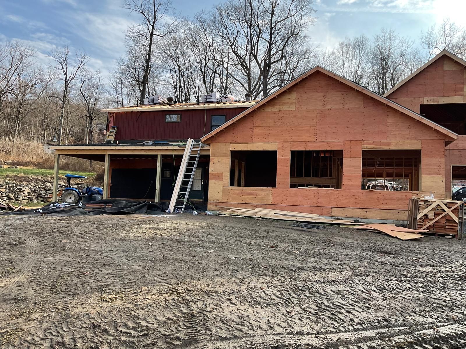 A house under construction with exposed wooden framing and siding, a ladder, and a gravel yard on a sunny day.