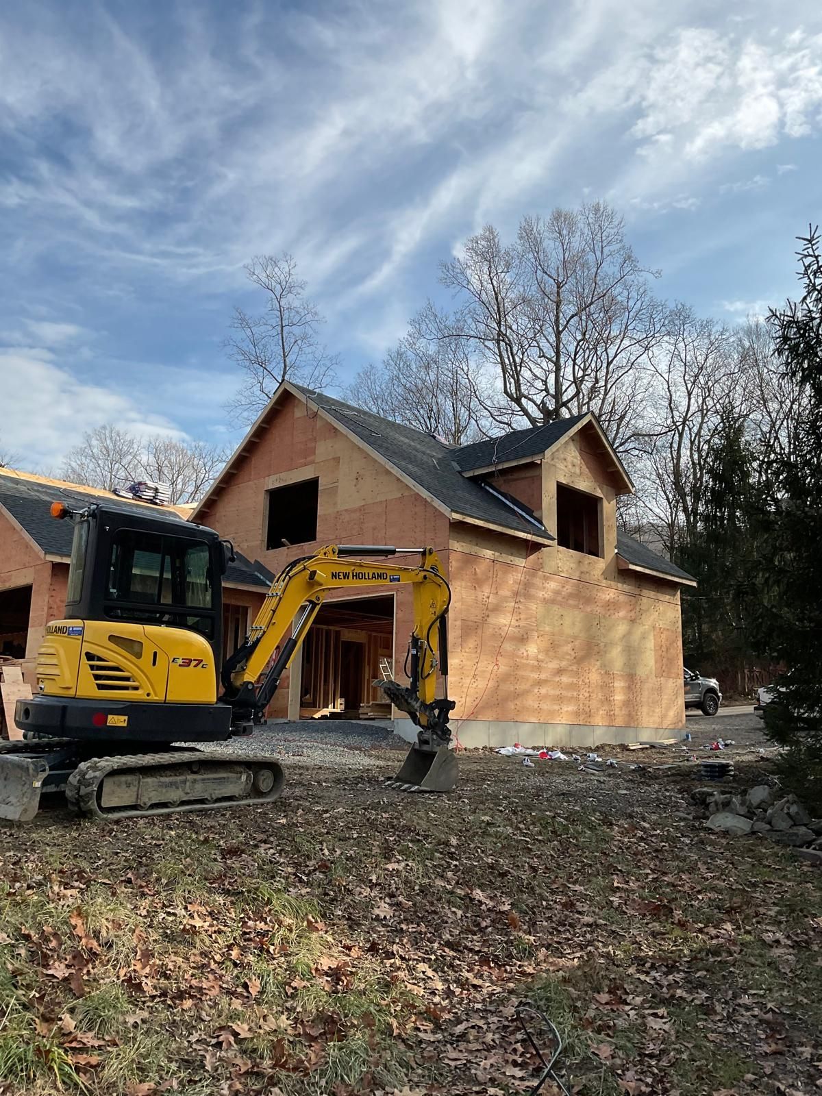 A yellow mini excavator parked in front of a wooden house frame under construction against a cloudy blue sky.