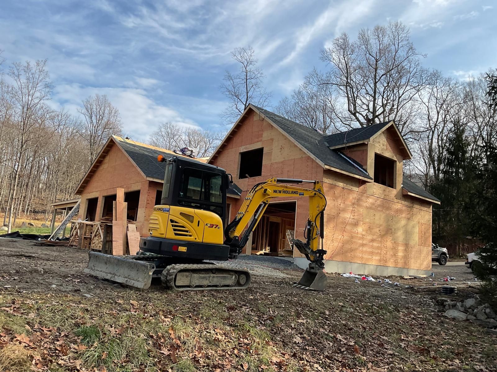 A yellow excavator parked in front of a partially constructed wooden house under a cloudy sky.
