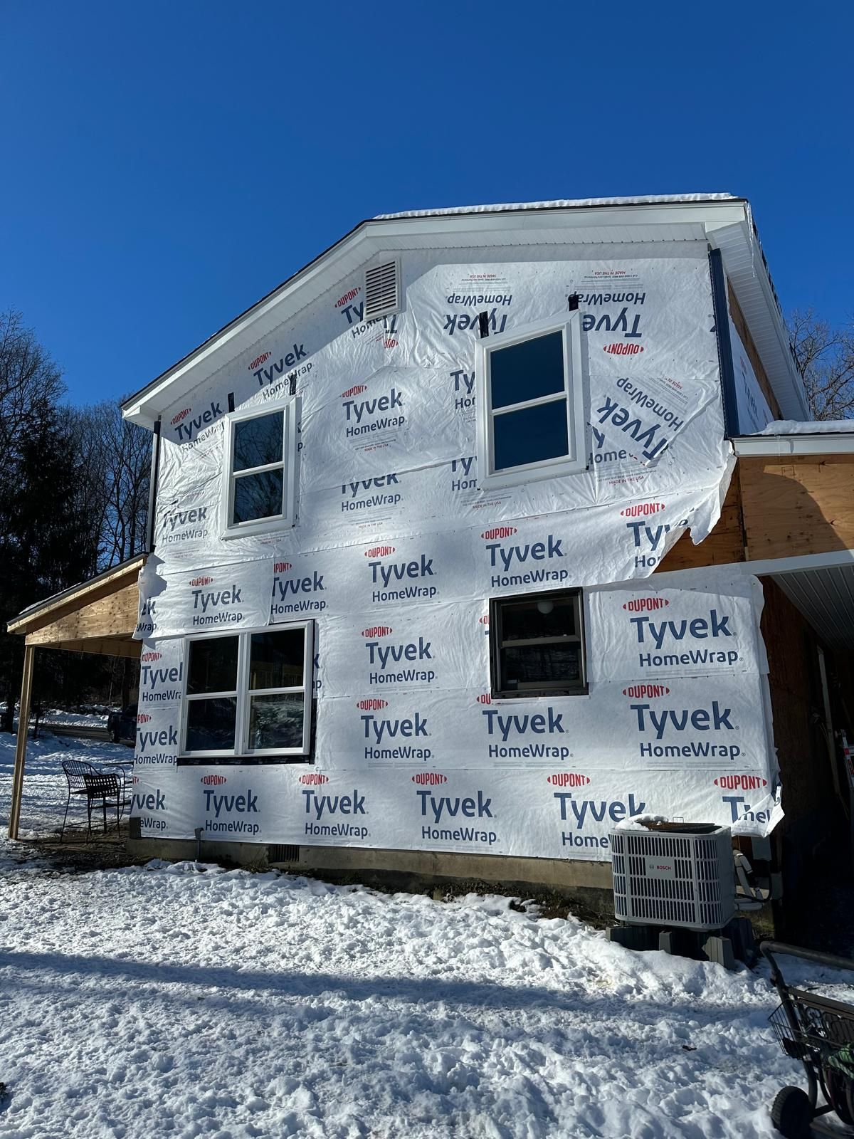 A house under construction wrapped in white Tyvek weather barrier with installed windows on a snowy day.