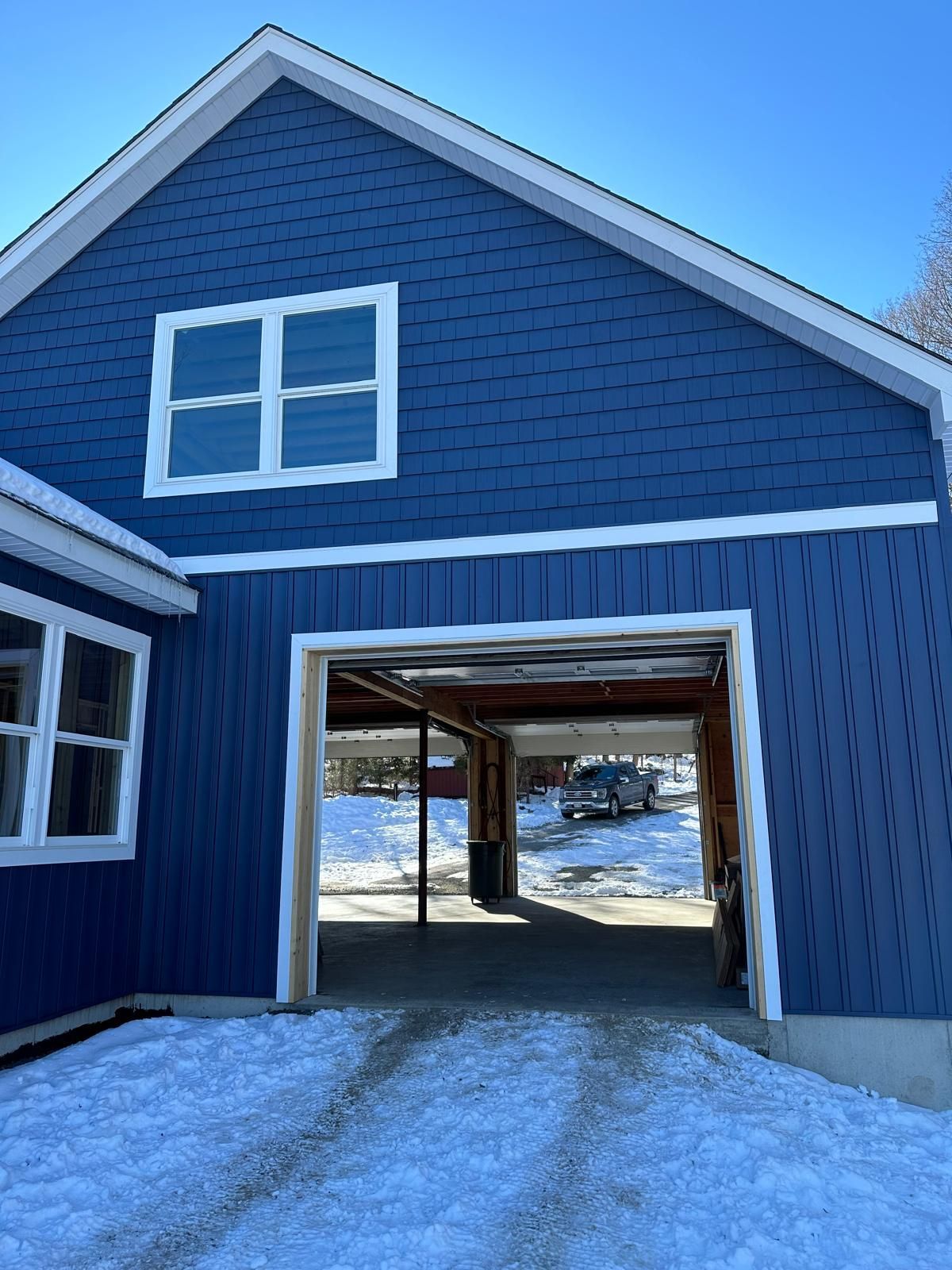 A two-story blue building with white trim featuring an open-air garage bay on the ground level, surrounded by snow.