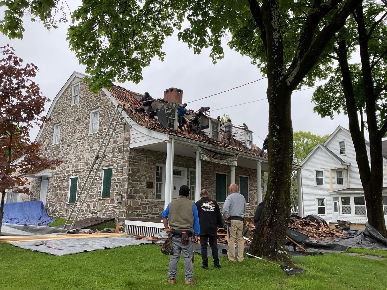 Three people stand on a lawn watching workers remove a damaged roof from a two-story stone house under a cloudy sky.