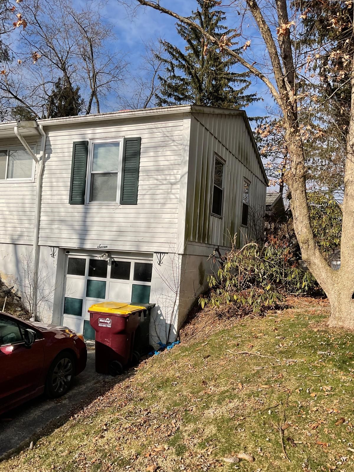 A two-story white house with a garage, dark green shutters, and a yellow-lidded trash bin parked on a sloped driveway.