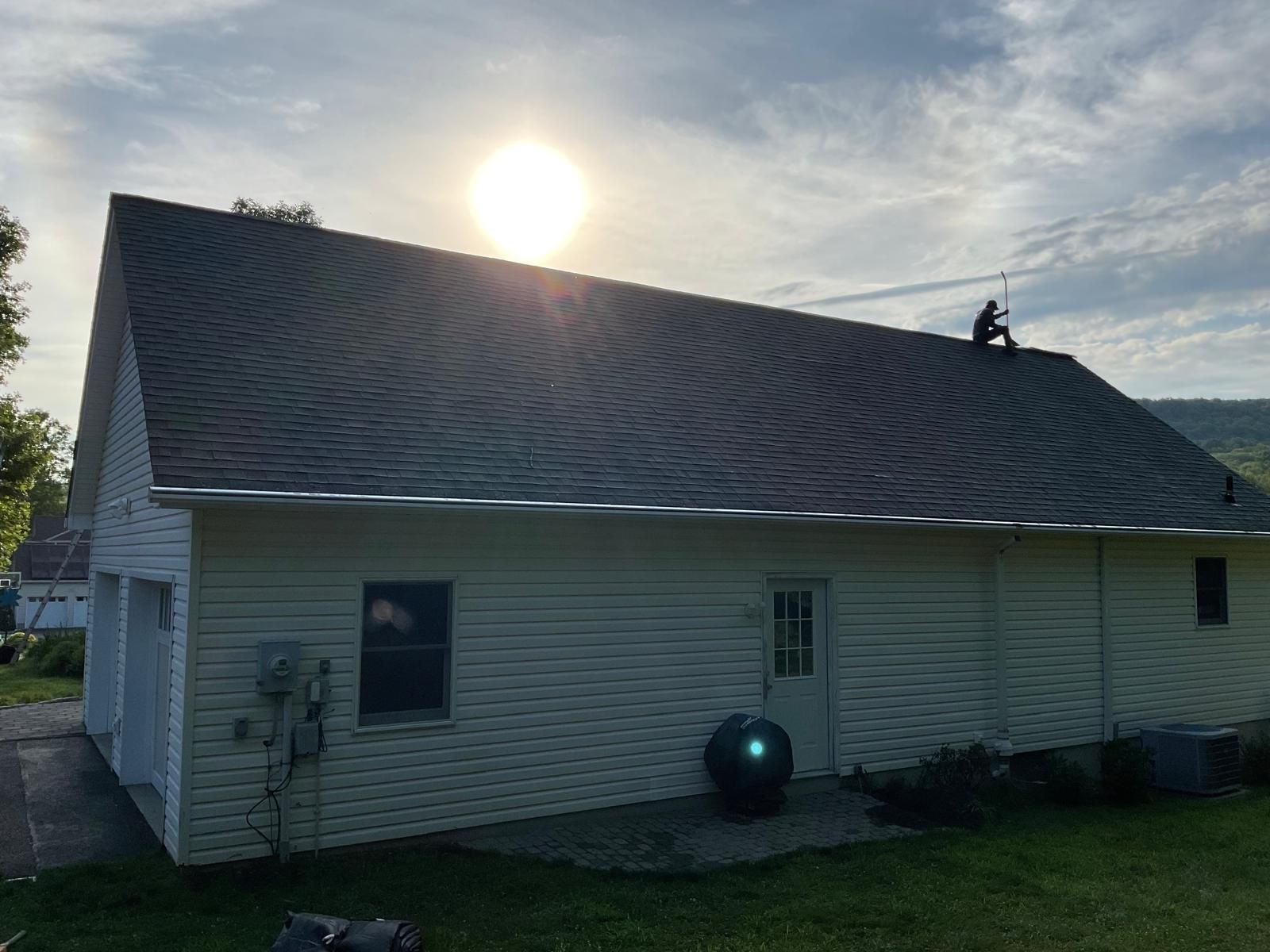 A person sits atop the pitched shingled roof of a light-sided suburban house at sunset.