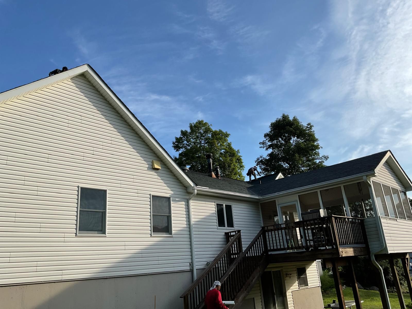 Two people work on the roof of a house with white siding, a wooden deck, and a staircase under a blue sky.