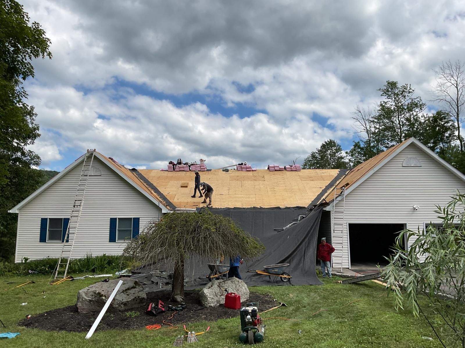 Workers installing new plywood sheathing on the roof of a house with white siding under a cloudy sky.