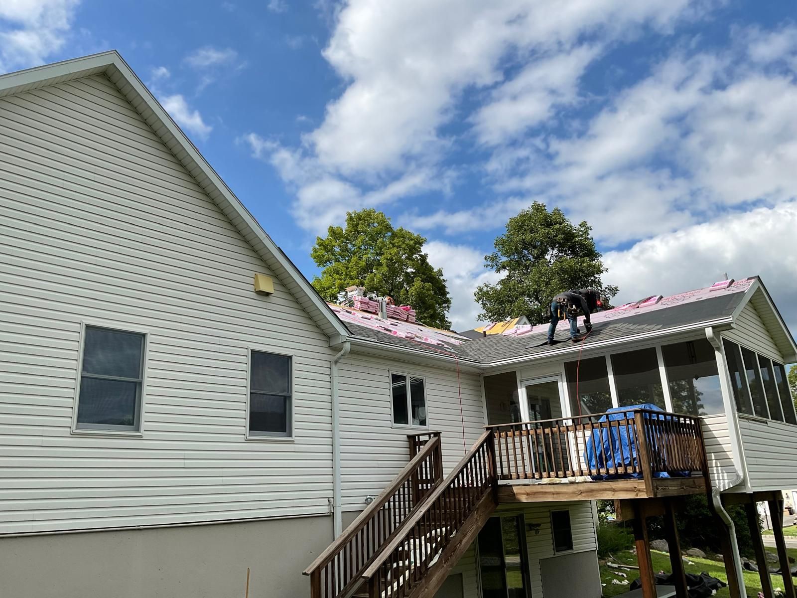 Workers install new shingles on the roof of a house with white siding and an attached deck under a partly cloudy sky.