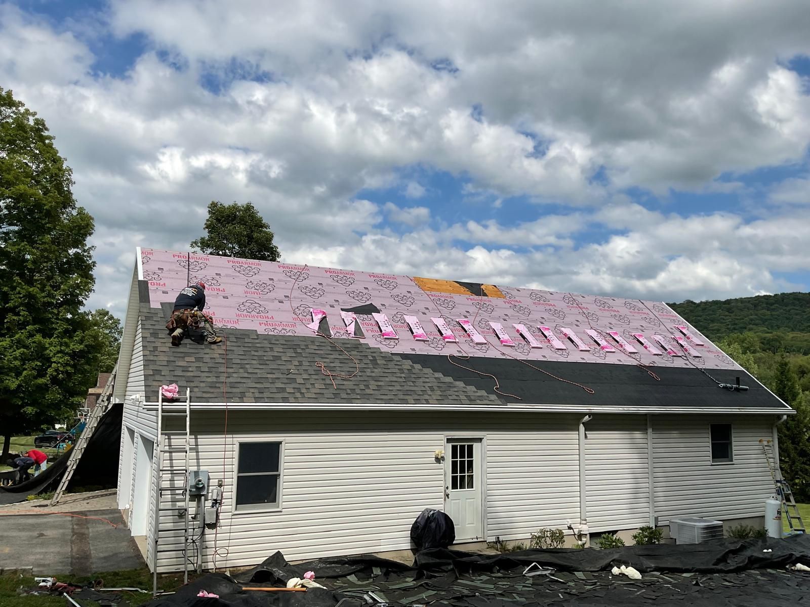 A construction worker on the roof of a house, installing dark gray shingles over pink foam board insulation.