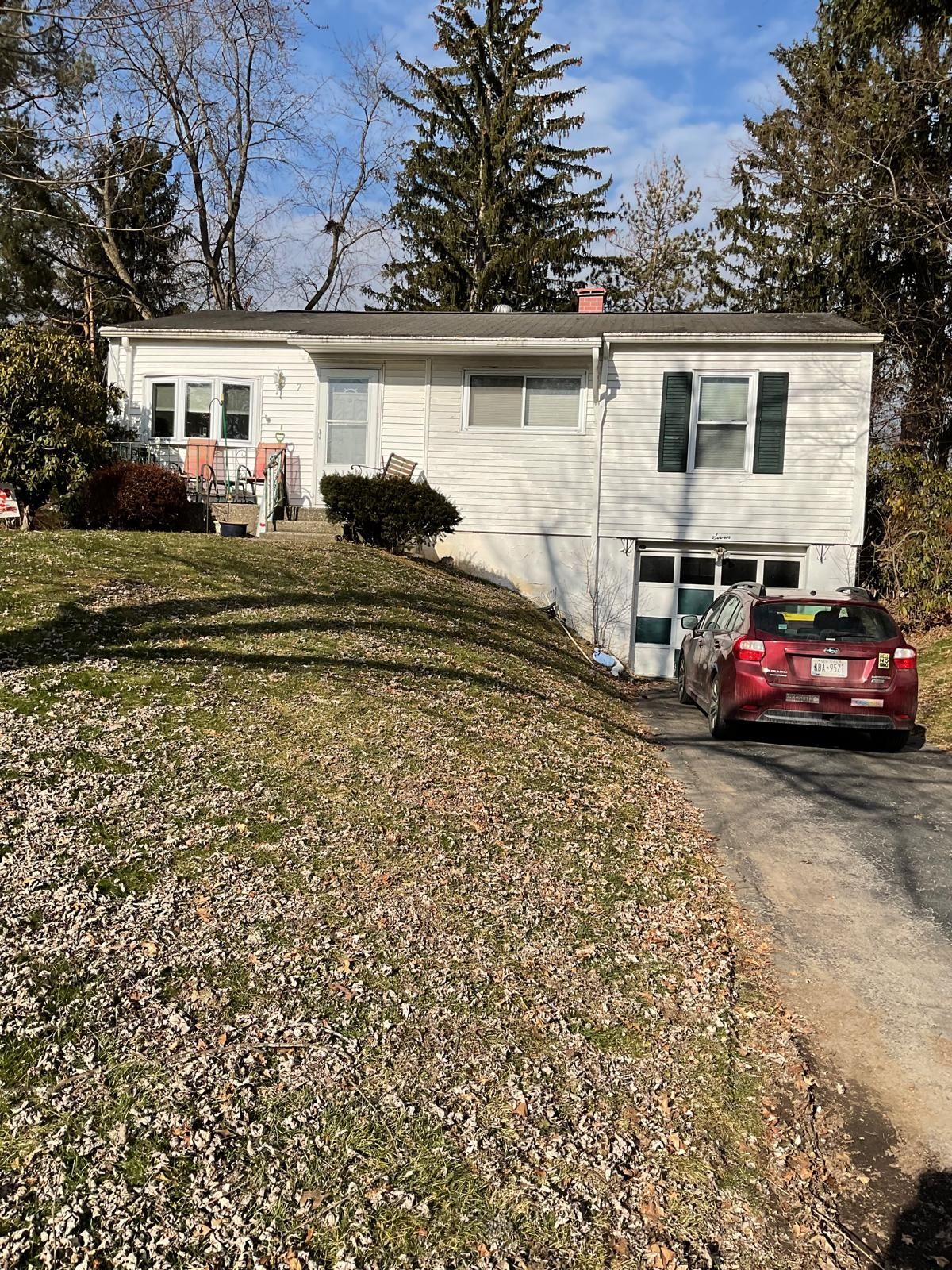 A white, single-story house with a basement garage and a sloped, rock-covered front yard, featuring a parked red car.
