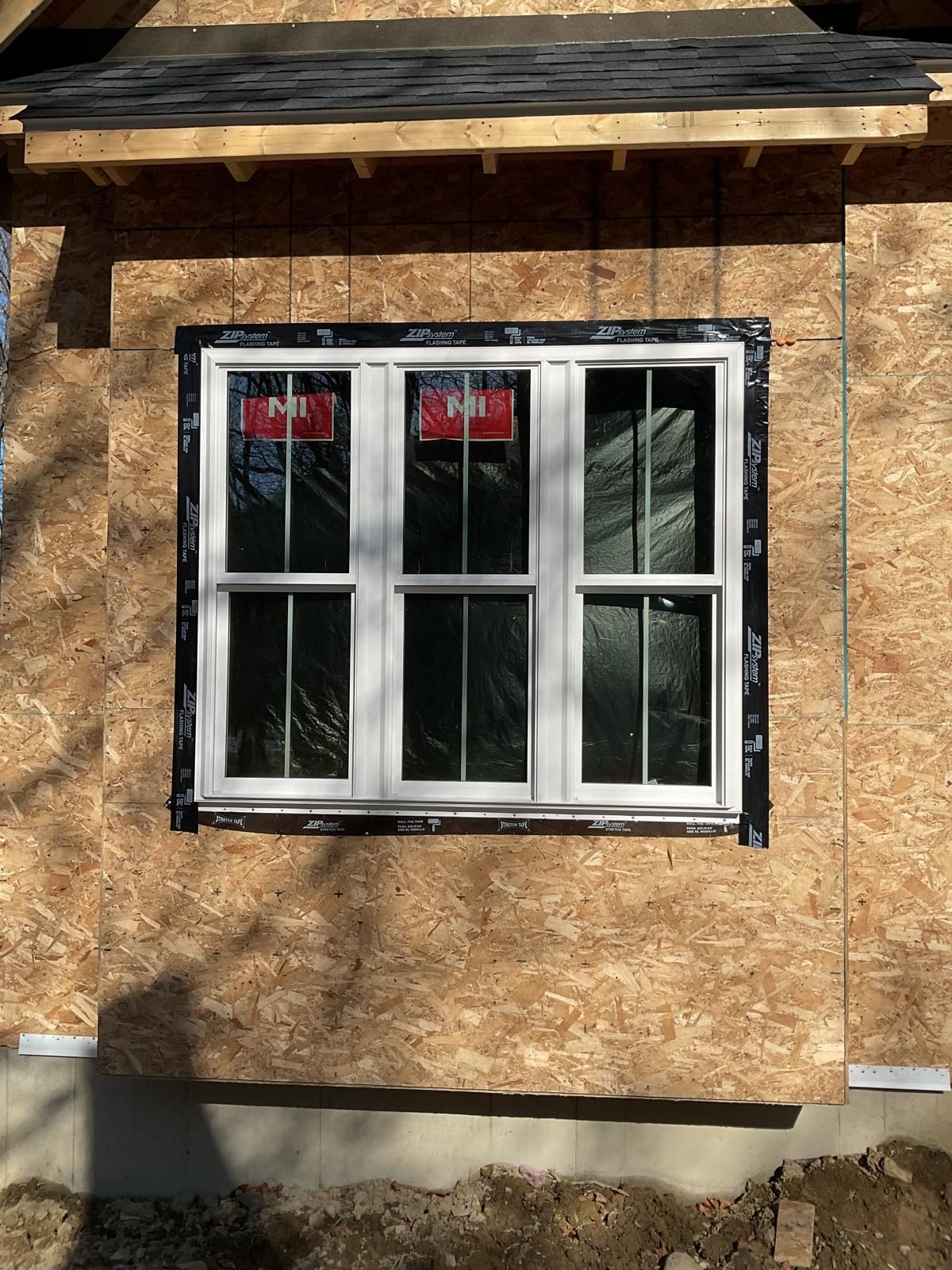 A three-pane white window installed in an exterior wall covered with oriented strand board (OSB) sheathing.