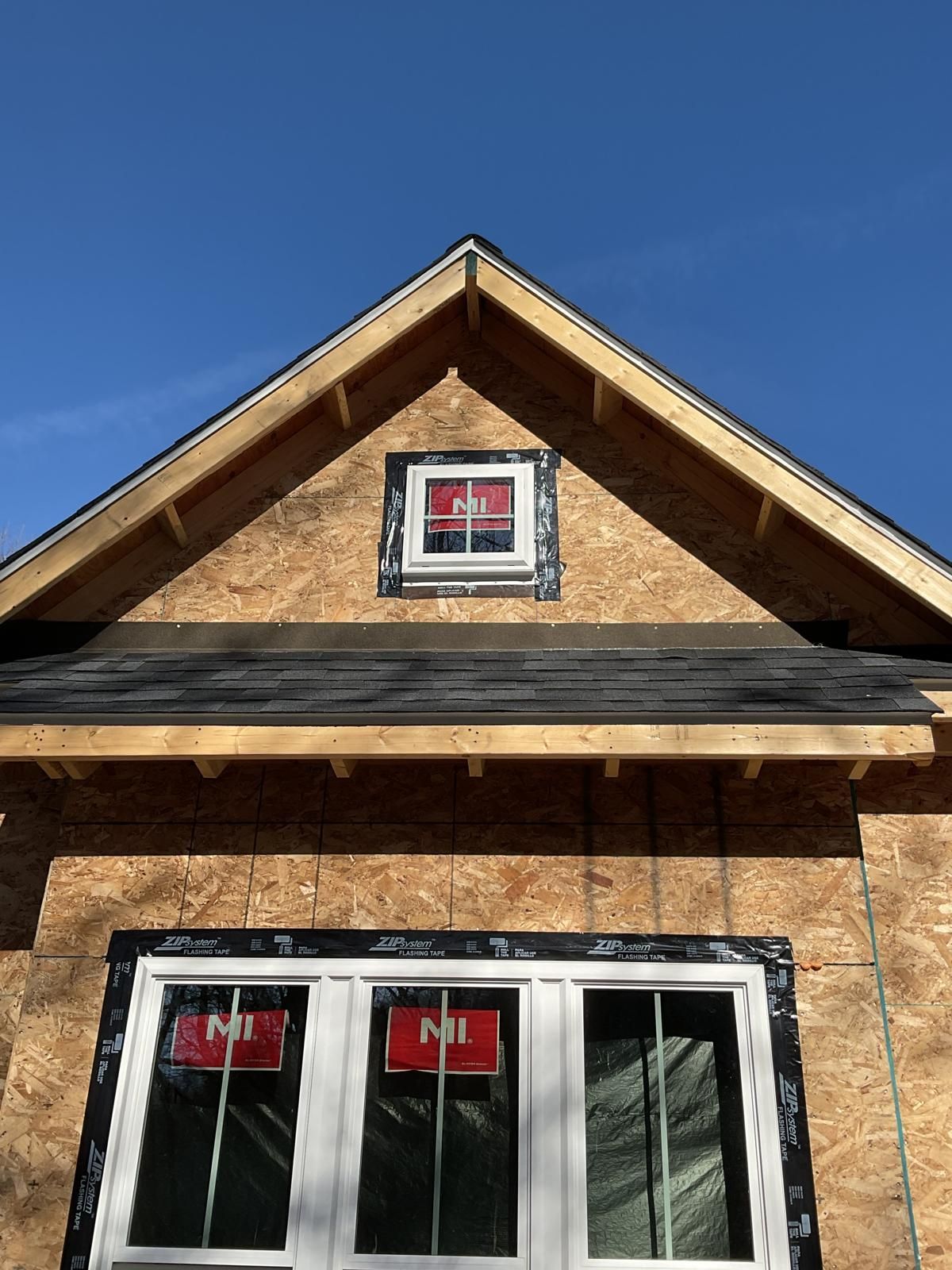 Construction exterior of a house under building, featuring a gabled roof, plywood siding, and installed windows.