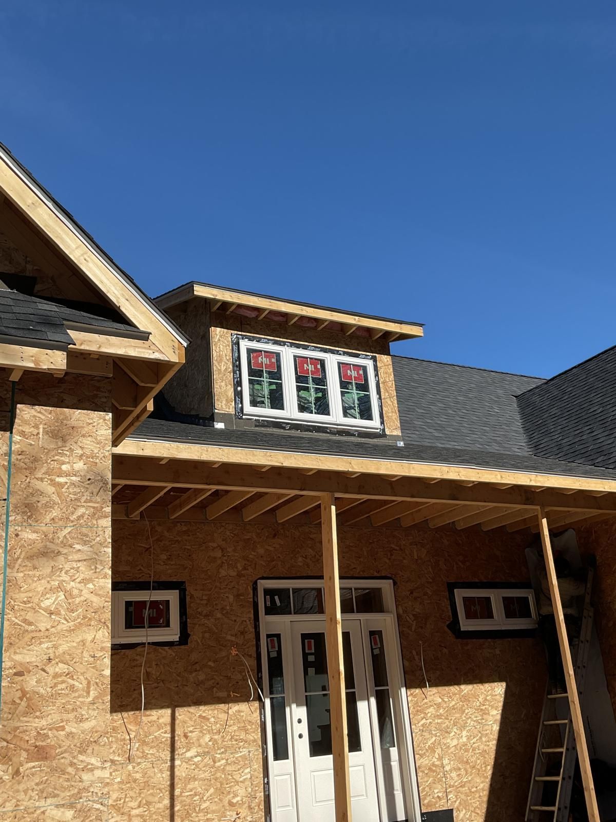 A house under construction showing a front entryway, a wooden porch frame, and an upper dormer window against a blue sky.