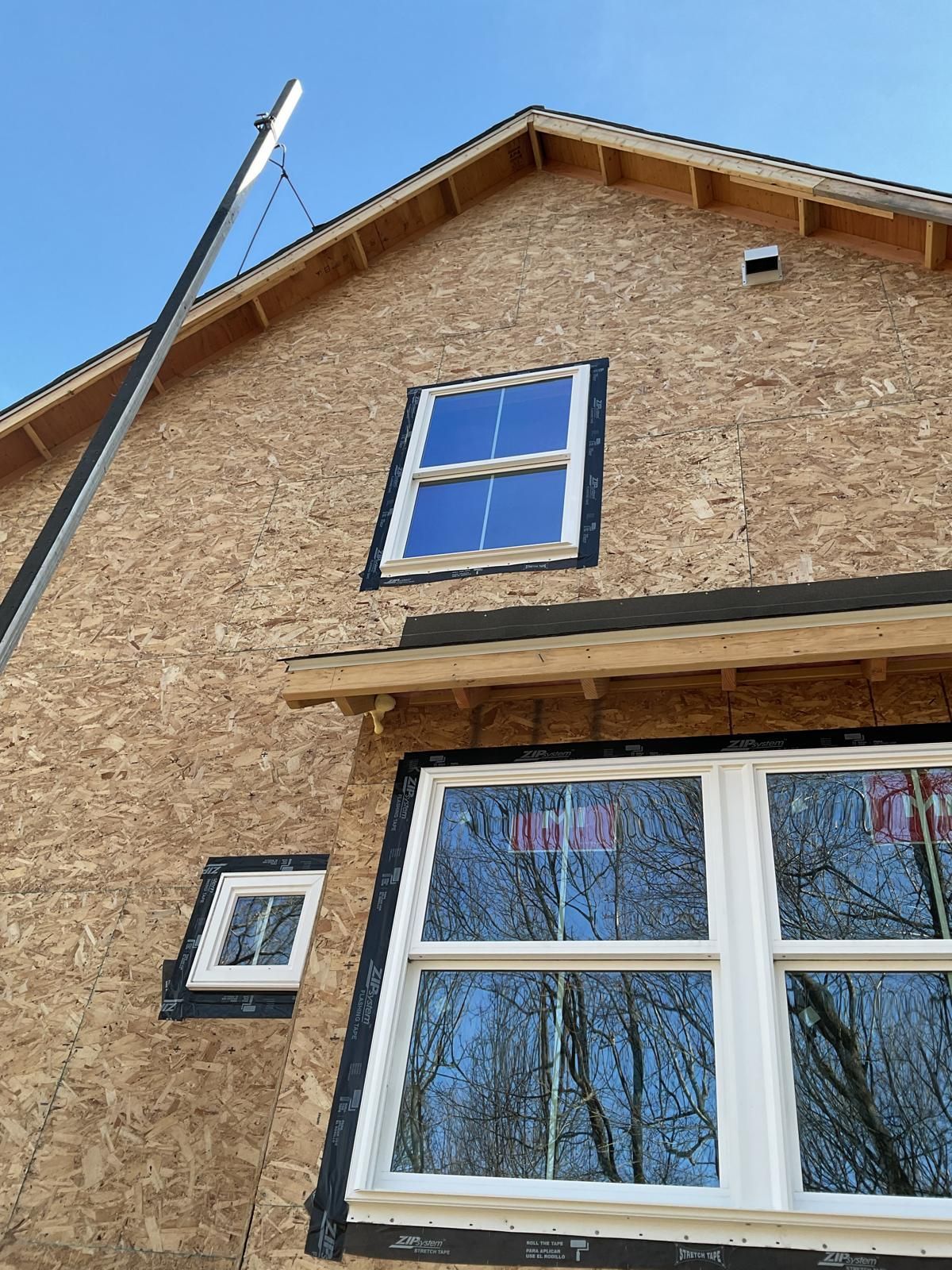 Exterior view of a house under construction with OSB sheathing and three newly installed windows against a blue sky.
