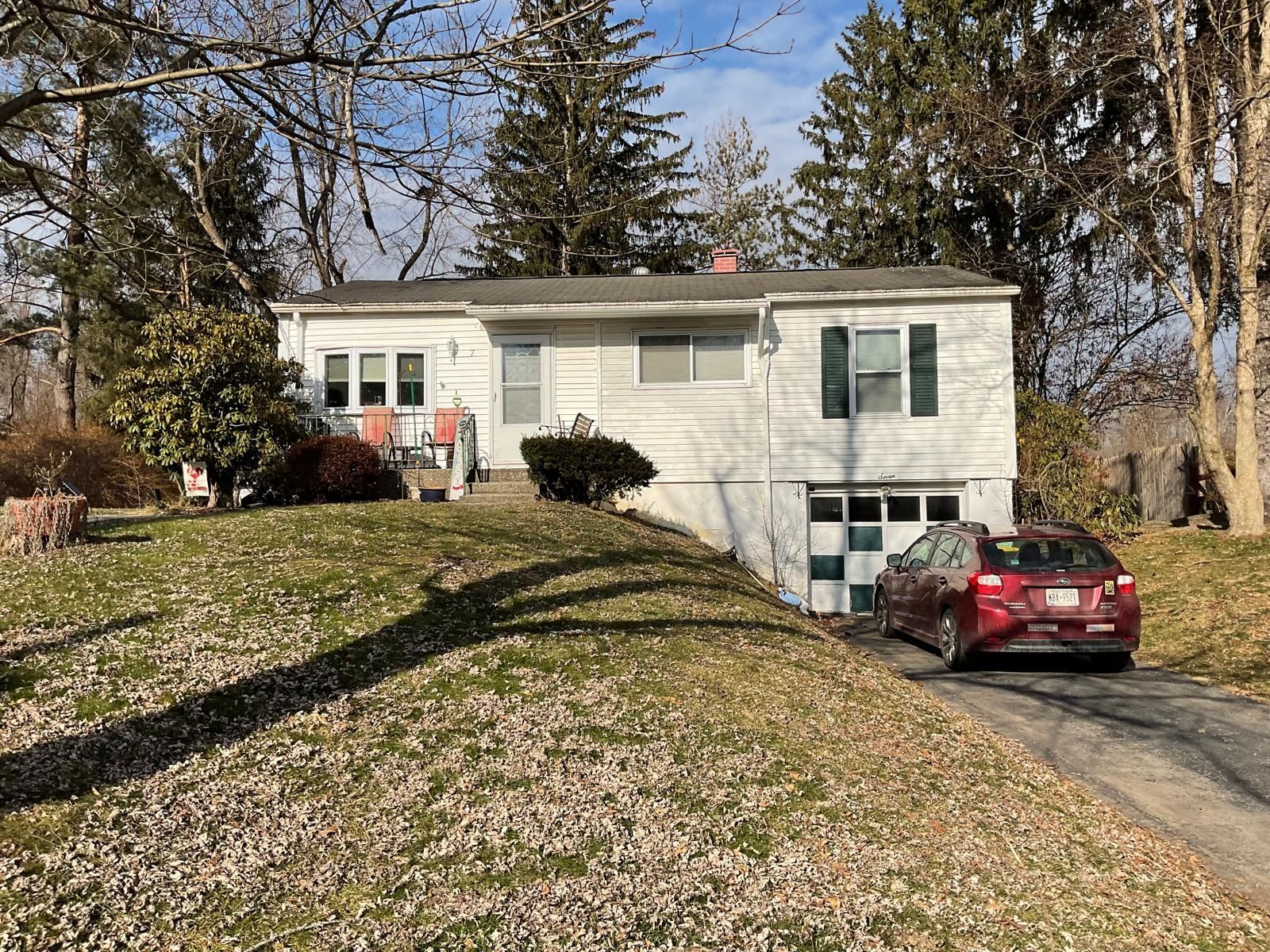 A white split-level house with a red car parked in the driveway, surrounded by trees and a sloped lawn in daylight.