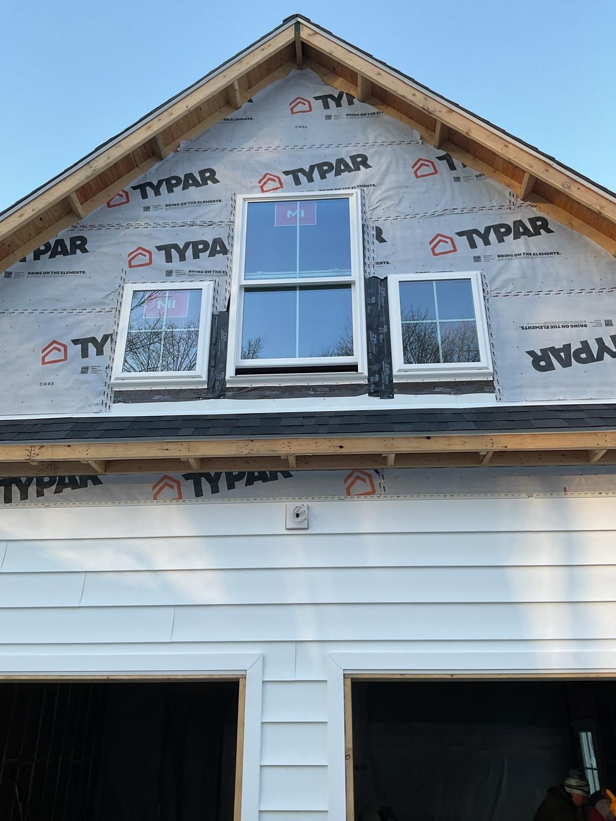 Exterior view of a gable roofline with Typar house wrap, three installed windows, and partially installed white siding.