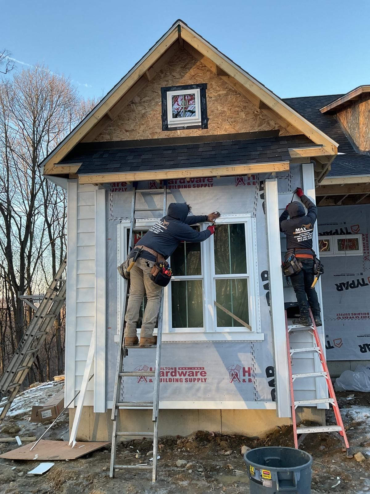 Two construction workers stand on ladders installing trim around windows on the exterior of a house under construction.