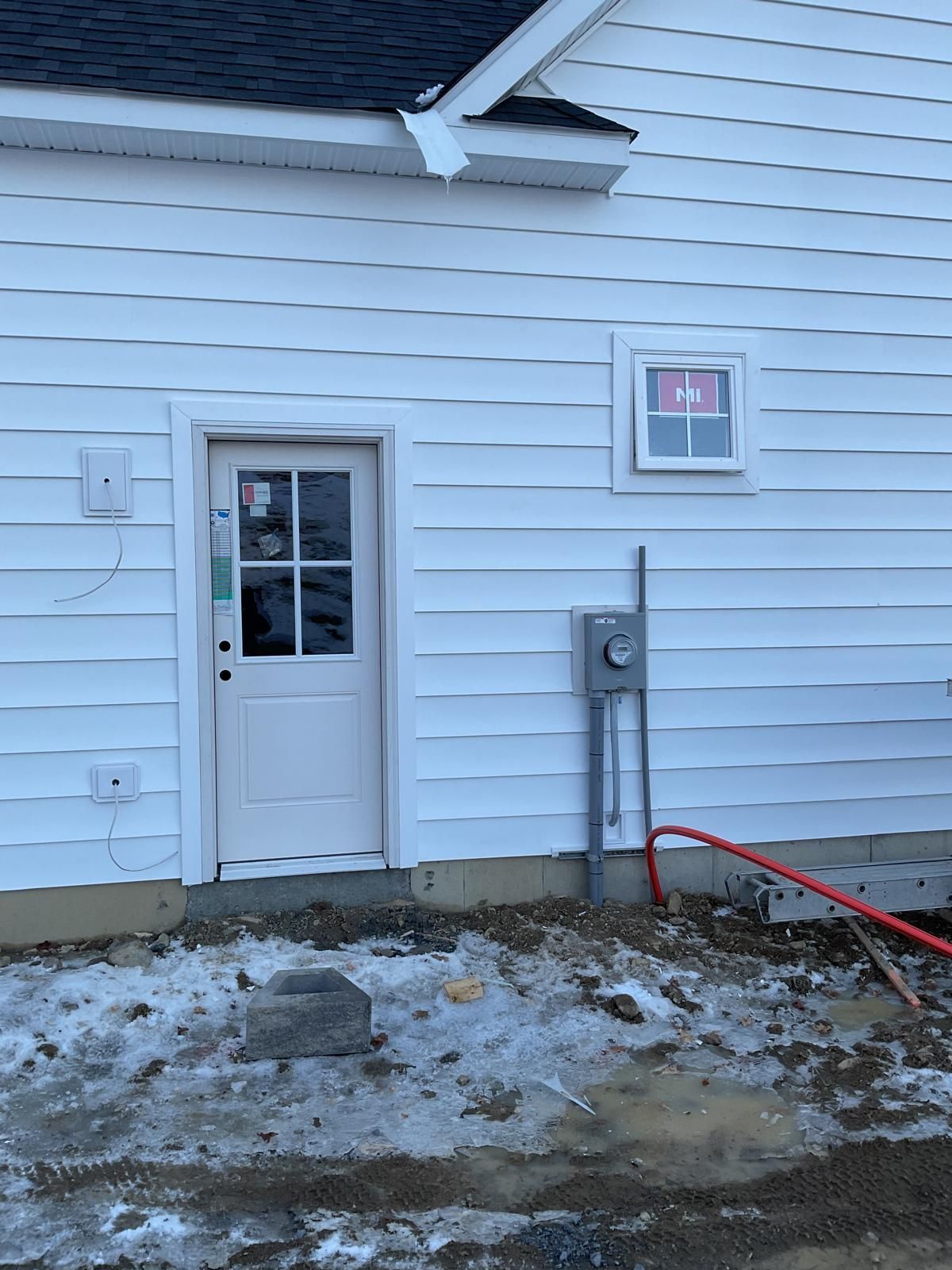 A white, vinyl-sided building exterior with a small door, a window, and an electrical meter on a snowy, muddy ground.
