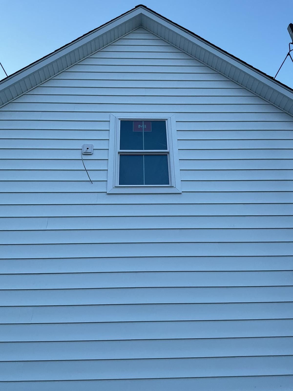 A white, horizontal-sided house gable with a centered, rectangular window and a small wall-mounted fixture to the left.