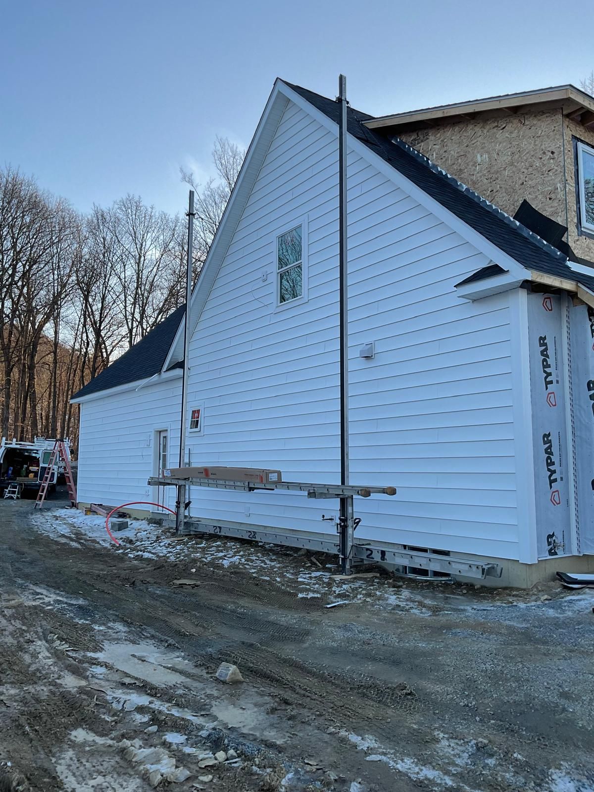 A new two-story house under construction with white siding, a dark roof, and scaffolding in a wooded lot.