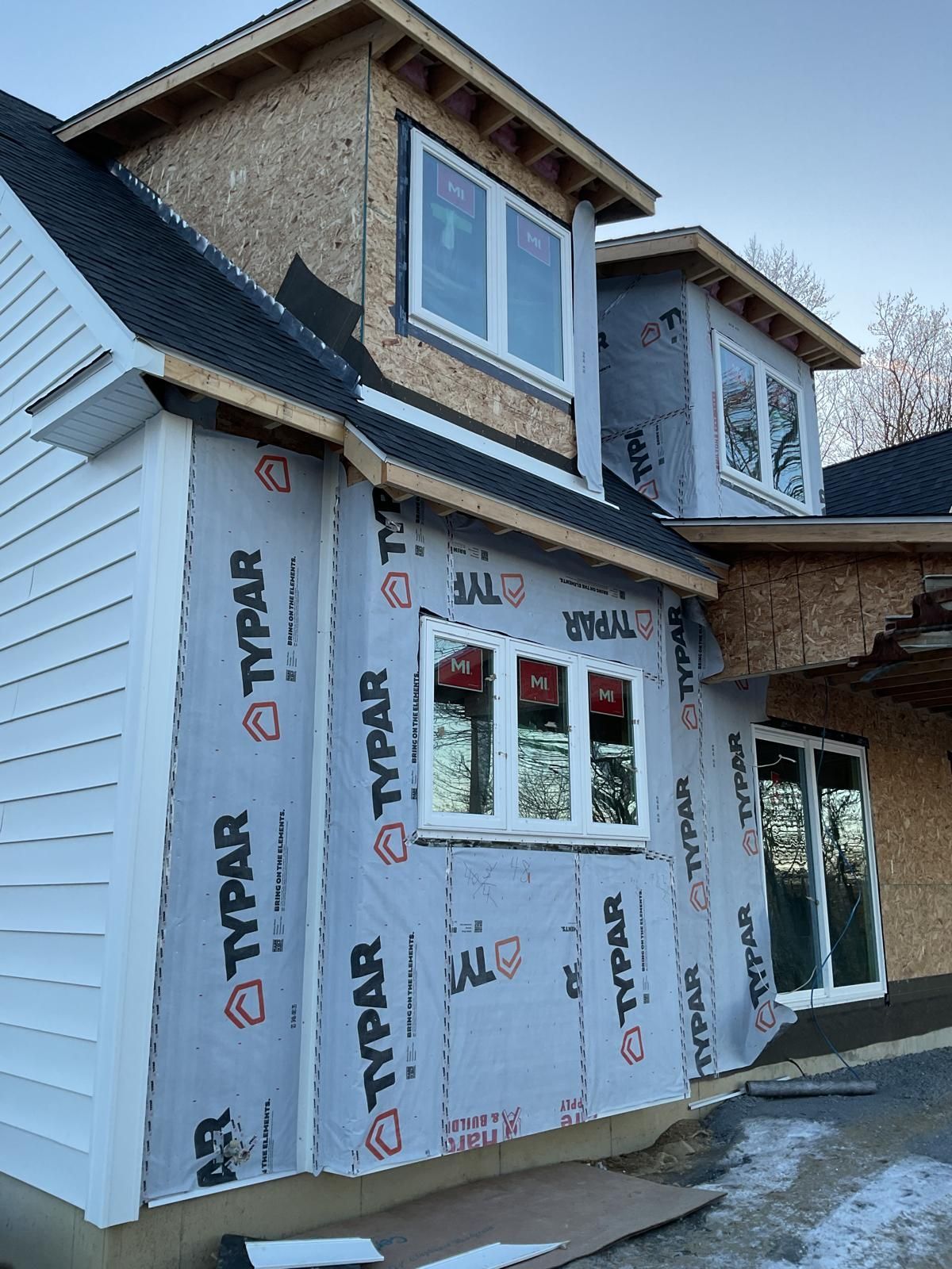 A house under construction with white siding, exposed plywood, and grey Typar weather-resistant barrier on the walls.